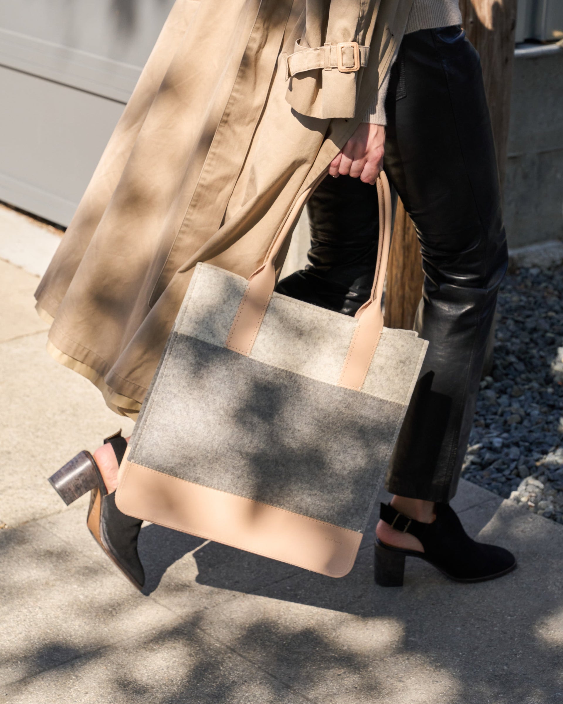 A granite-colored Jaunt Merino Wool Felt Tote bag is elegantly held by its top handle as a woman walks, showcasing its chic design