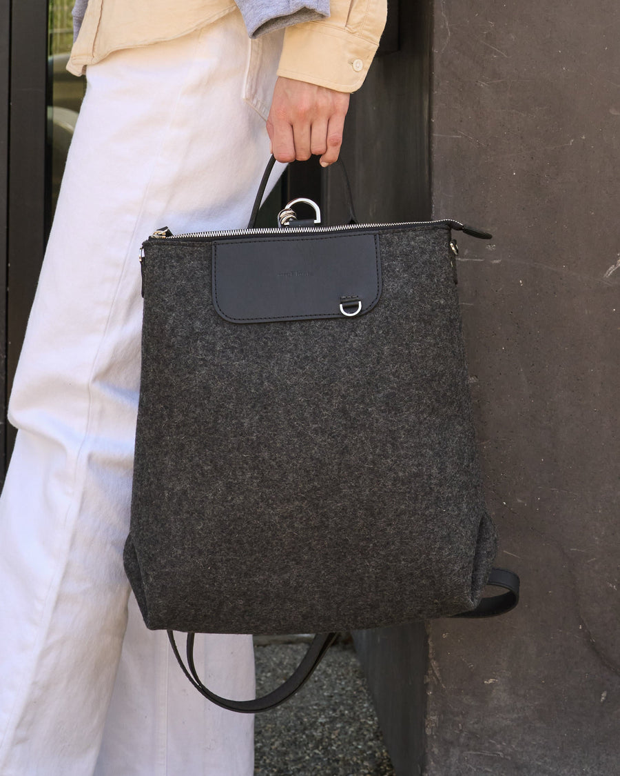 A charcoal-colored Bedford Merino Wool Felt Backpack held by the top handle on a woman's side while standing, highlighting its chic design
