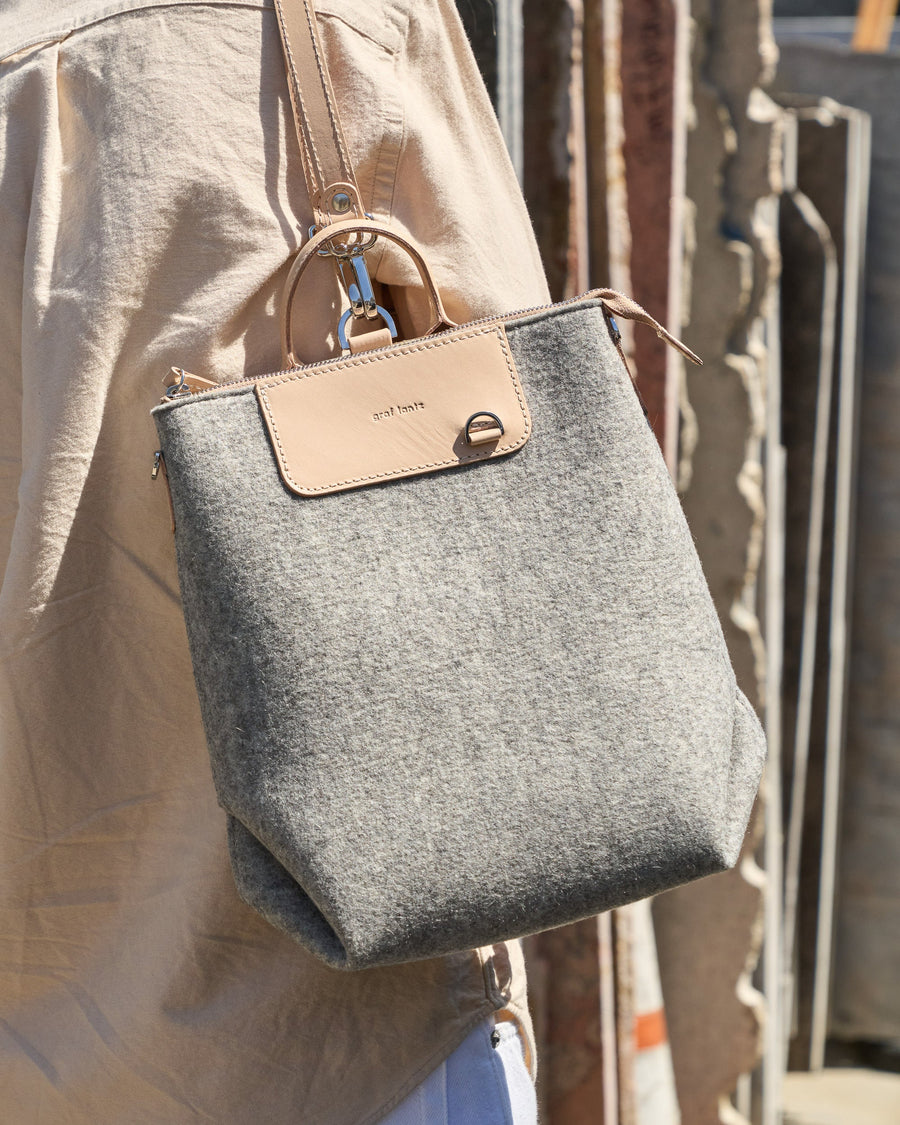 A Bedford Merino Wool Felt Midi Backpack carried by a woman over her shoulder, showcasing its light grey and beige colors
