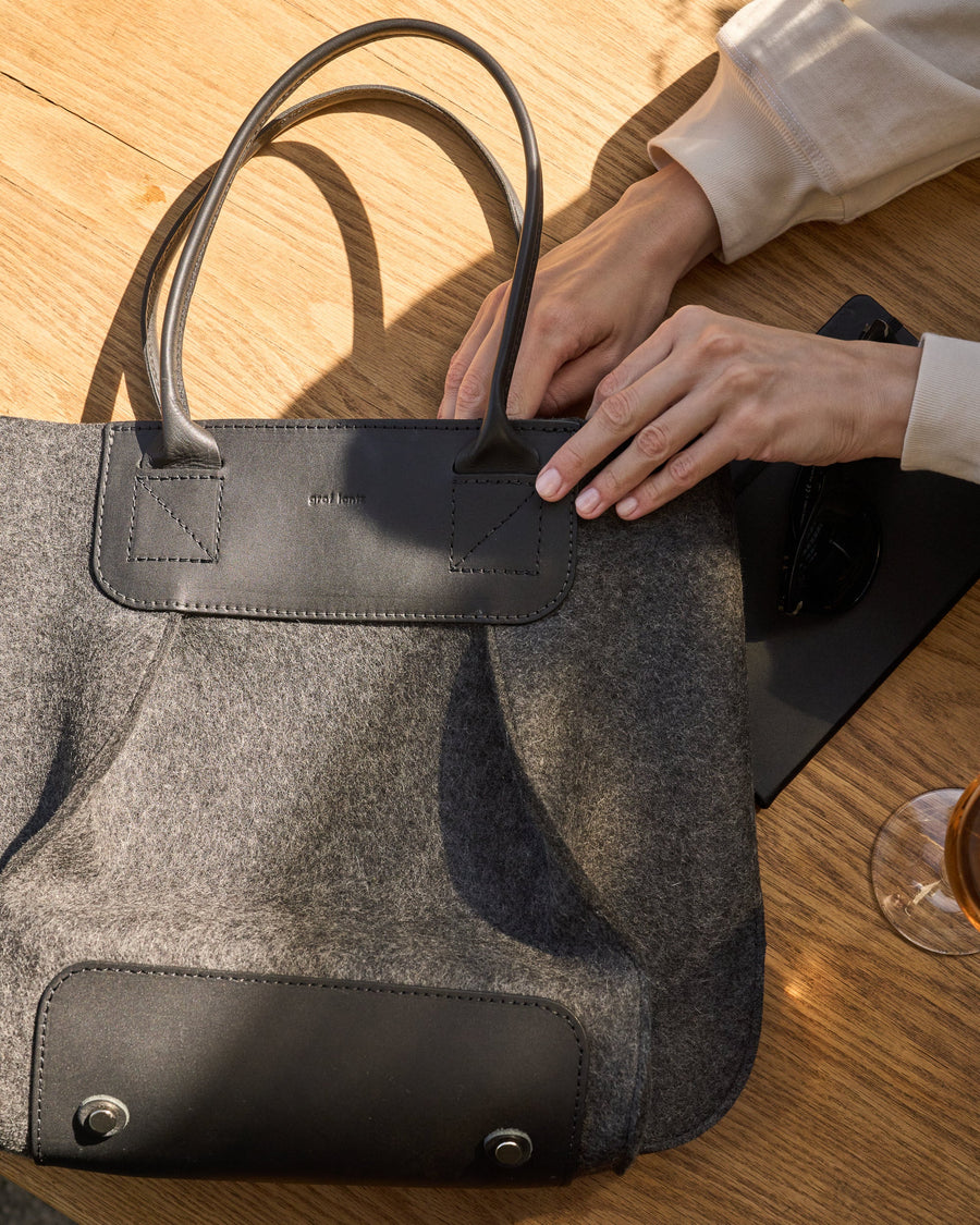 Hands retrieving something from the inside of a charcoal-colored Frankie Merino Wool Felt Midi tote bag placed on a wooden table beside a wine glass