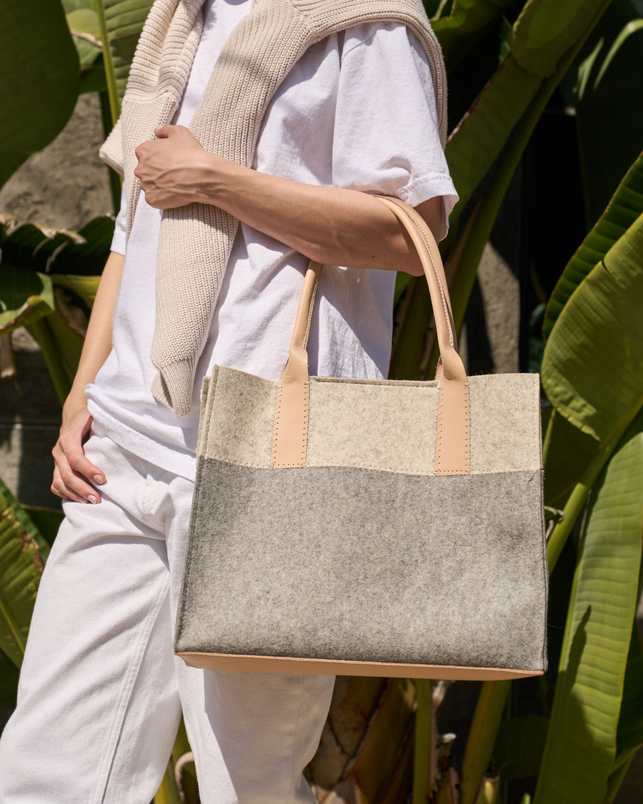 A woman carries a grey, white, and beige colored Jaunt Midi Merino Wool Felt Tote bag over her arm while walking, showcasing its medium size and timeless design