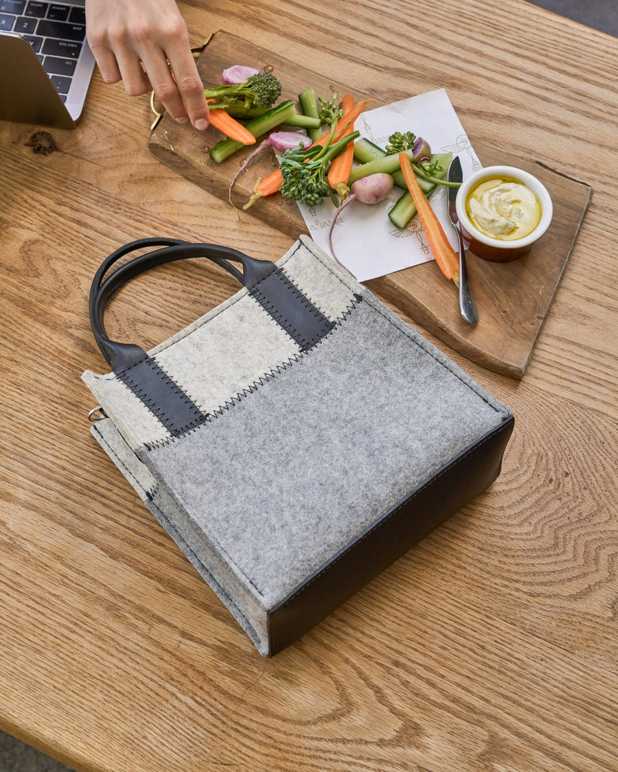 A granite and white-colored Jaunt Mini Merino Wool Felt Tote is placed on a wooden table next to a laptop and a tray with vegetable finger food