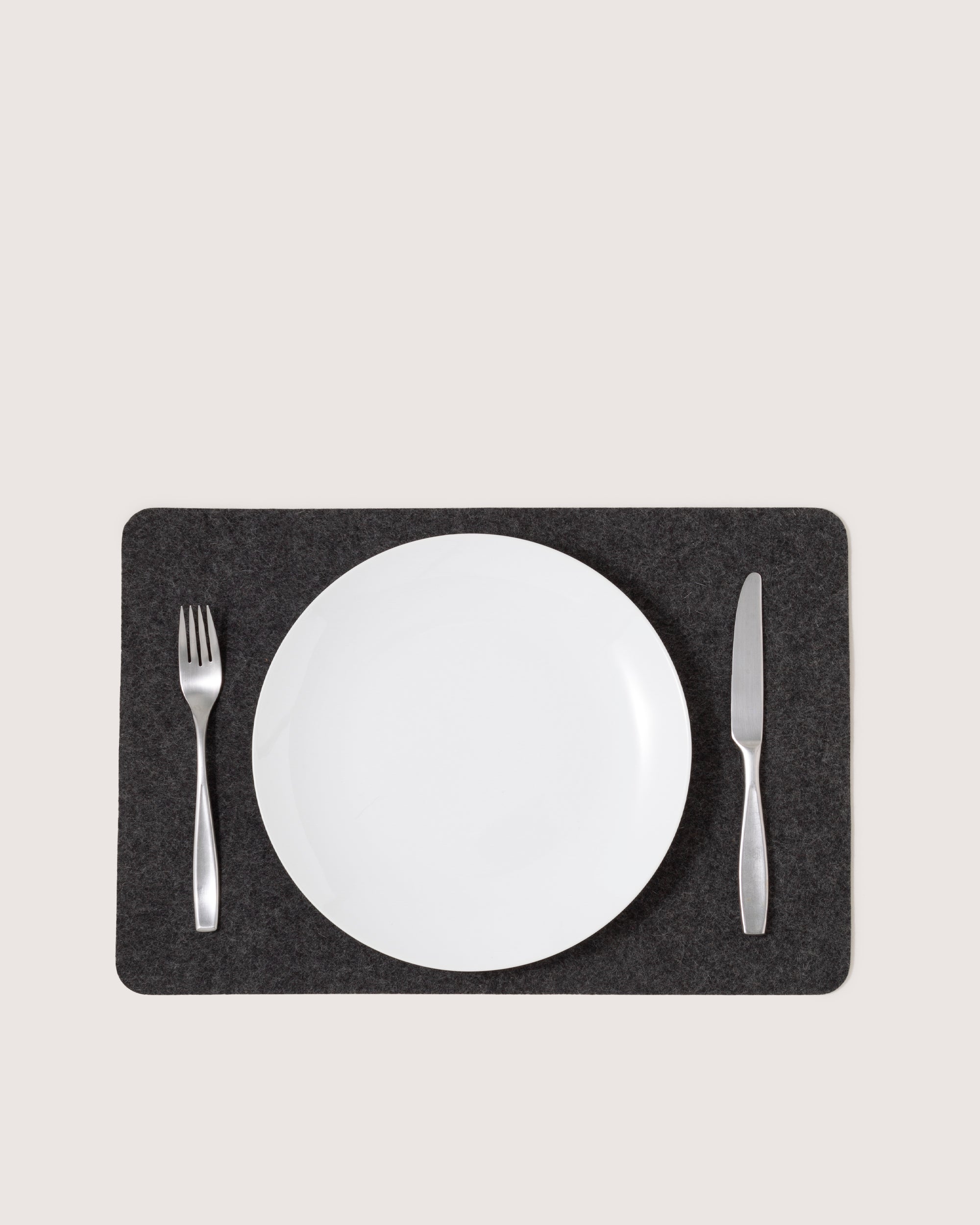 A white plate is centered on a dark graf lantz Merino Wool Placemat, known for its moisture-wicking properties, with a silver fork to the left and a silver knife to the right against a plain light background.