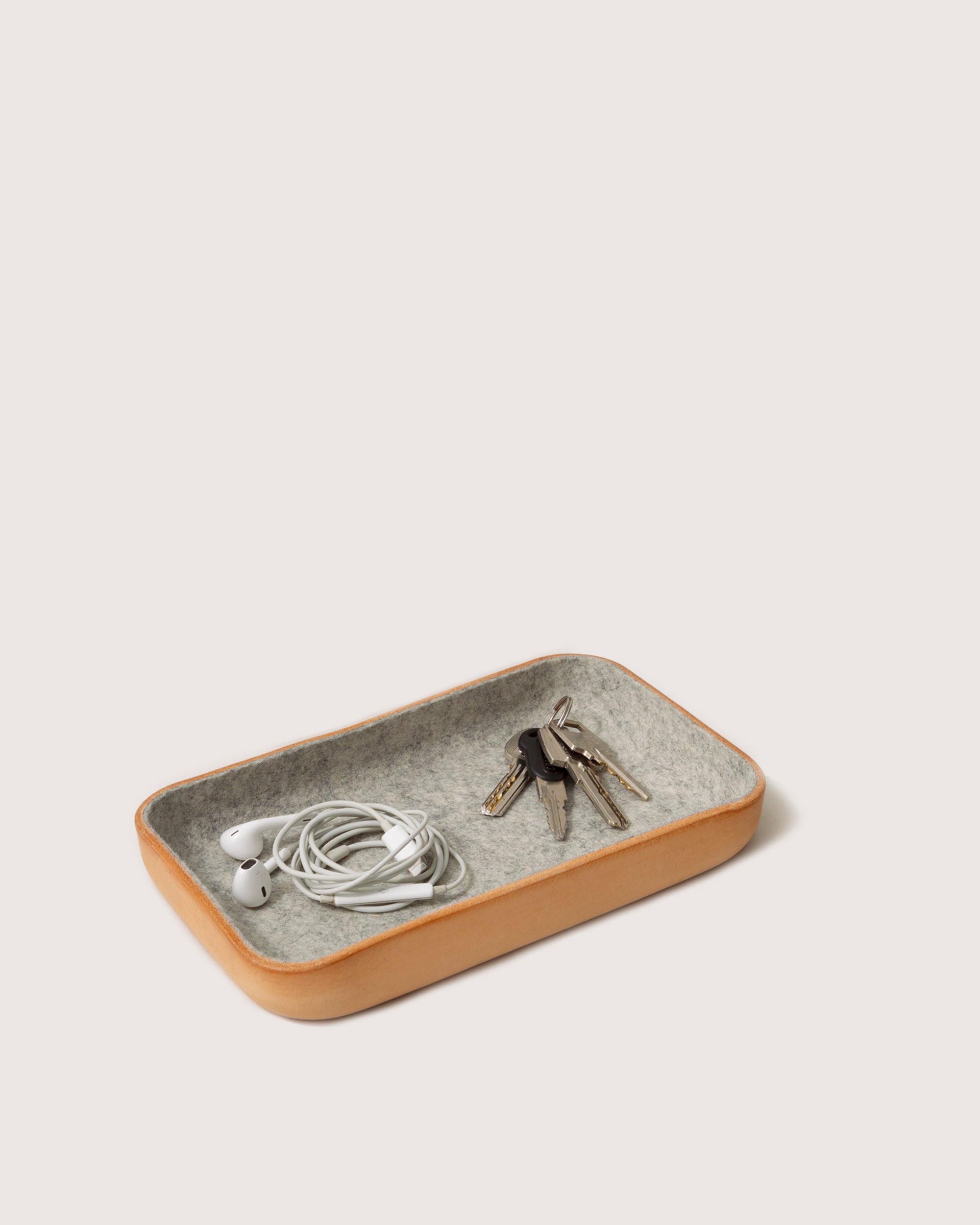 The graf lantz Kawabon Tray, made of Vachetta leather with a felt interior, elegantly organizes white earphones on the left and keys on the right. Its light brown exterior offers a stunning contrast against a plain white background.