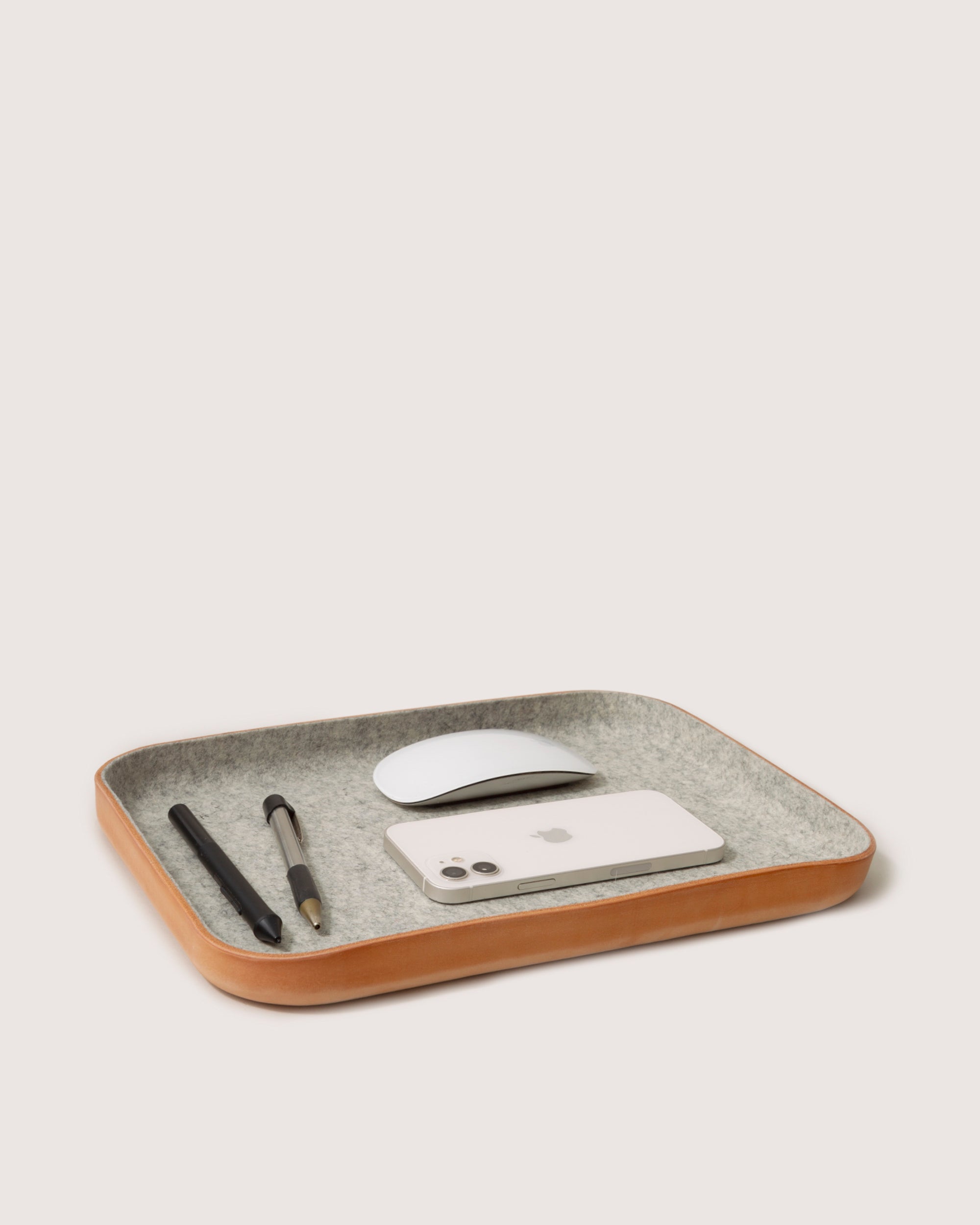 The graf lantz Kawabon Tray is a rectangular desk organizer made from Merino wool and Vachetta leather, elegantly holding a smartphone, wireless mouse, pen, and pencil in its minimalist design.