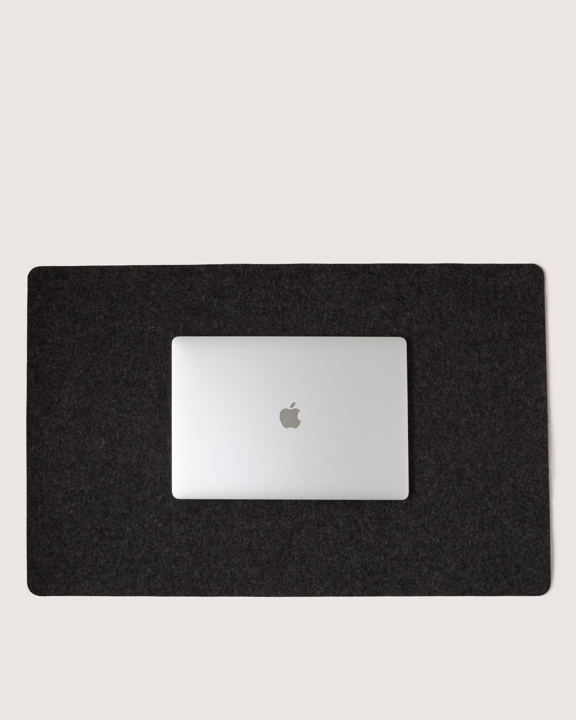 A closed silver laptop with an Apple logo sits on a graf lantz Mosen Desk Pad made of Merino Wool, centered on a large black rectangular mat against a plain light background.