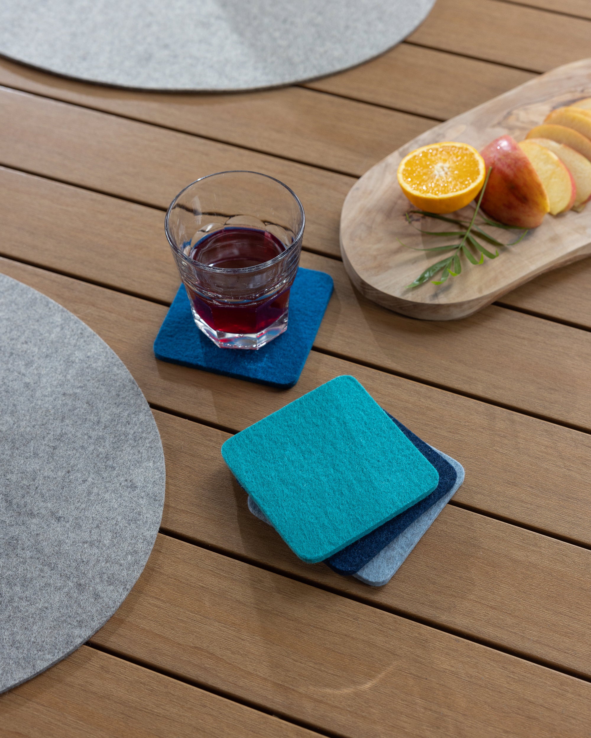 A glass of red liquid rests on a blue coaster on a wooden table. Nearby, a stack of graf lantz Beer Felt coasters, an oval tray with sliced apples and an orange half, and a gray placemat create an inviting scene.