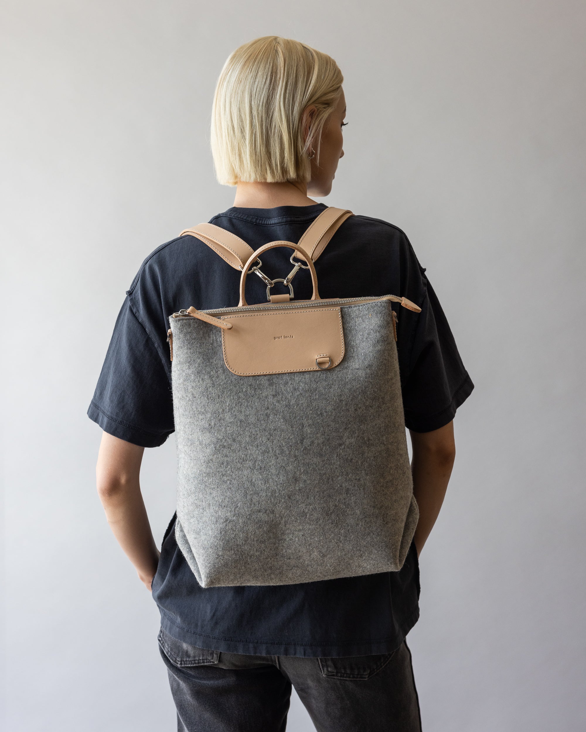 A person with blonde hair, facing away and wearing a black shirt and jeans, carries a graf lantz gray and beige backpack. Made of durable materials, the backpack boasts leather straps and a minimalist design, blending style with sustainability against a neutral background.