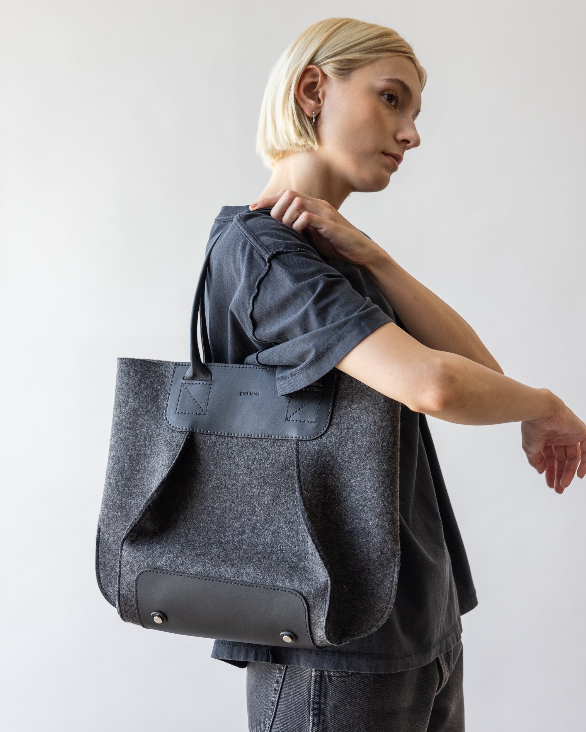 Someone with short blonde hair models a graf lantz Frankie Midi tote bag, featuring leather accents, metal studs, and a magnetic closure over their shoulder. The large gray tote contrasts sharply against the plain light-colored background.