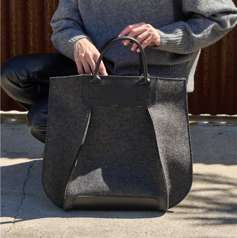 A Frankie Merino Wool Felt Tote bag held by a sitting woman, showcasing its dark grey and black colors