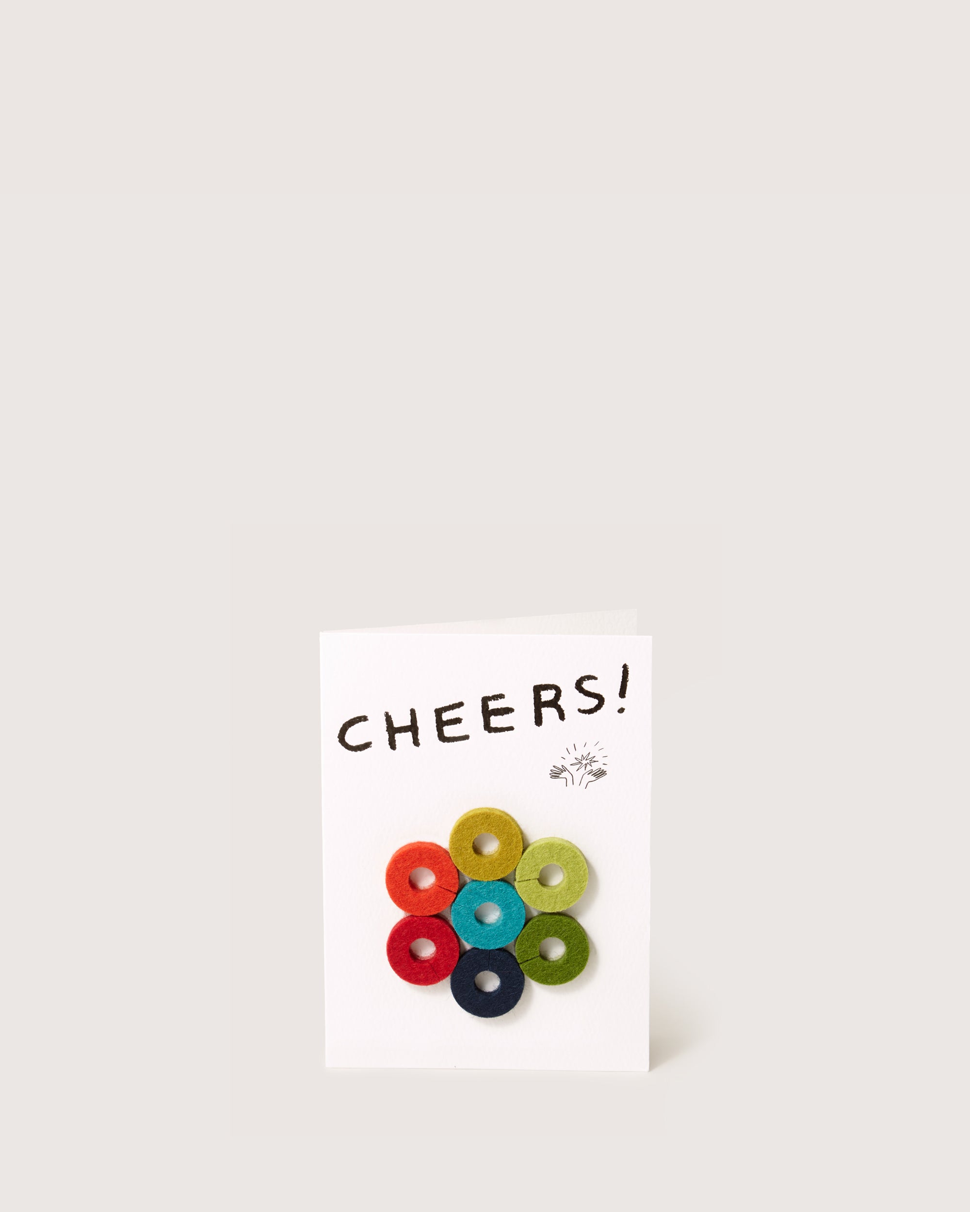 The graf lantz greeting card stands upright with CHEERS! at the top, showcasing seven colorful Merino wool felt circles in a floral design, making it an ideal host gift. It pops against its plain off-white background.
