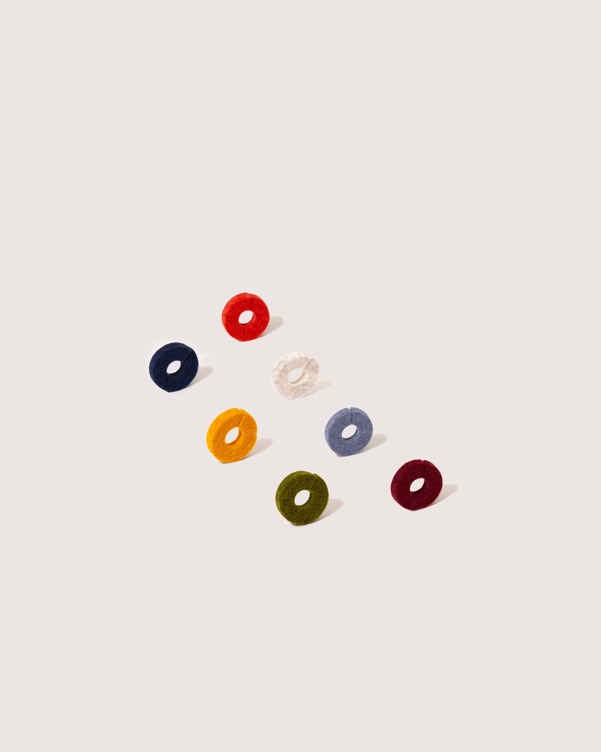 A perfect host gift, seven vibrant hair ties by graf lantz are arranged in a triangle on a light background. Made from Merino wool, they feature hues of navy blue, red, white, mustard, dusty blue, olive green, and maroon to add charm to any space.