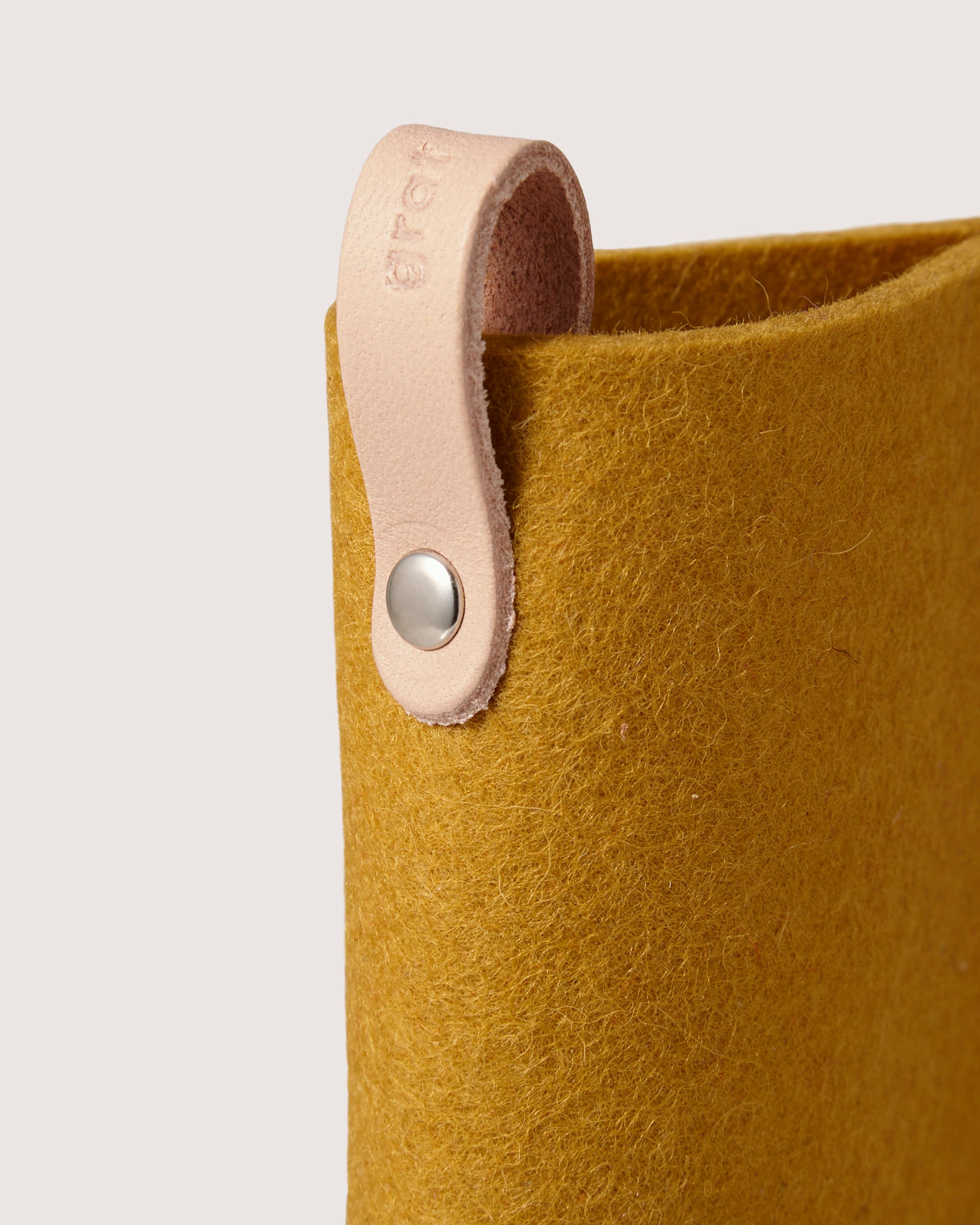 Close-up of a mustard felt storage basket by graf lantz, made from soft Merino wool. It features a light pink leather handle with metal button attachment in a minimalist design, perfect for holding an eyeglass sleeve or offering eyewear protection.