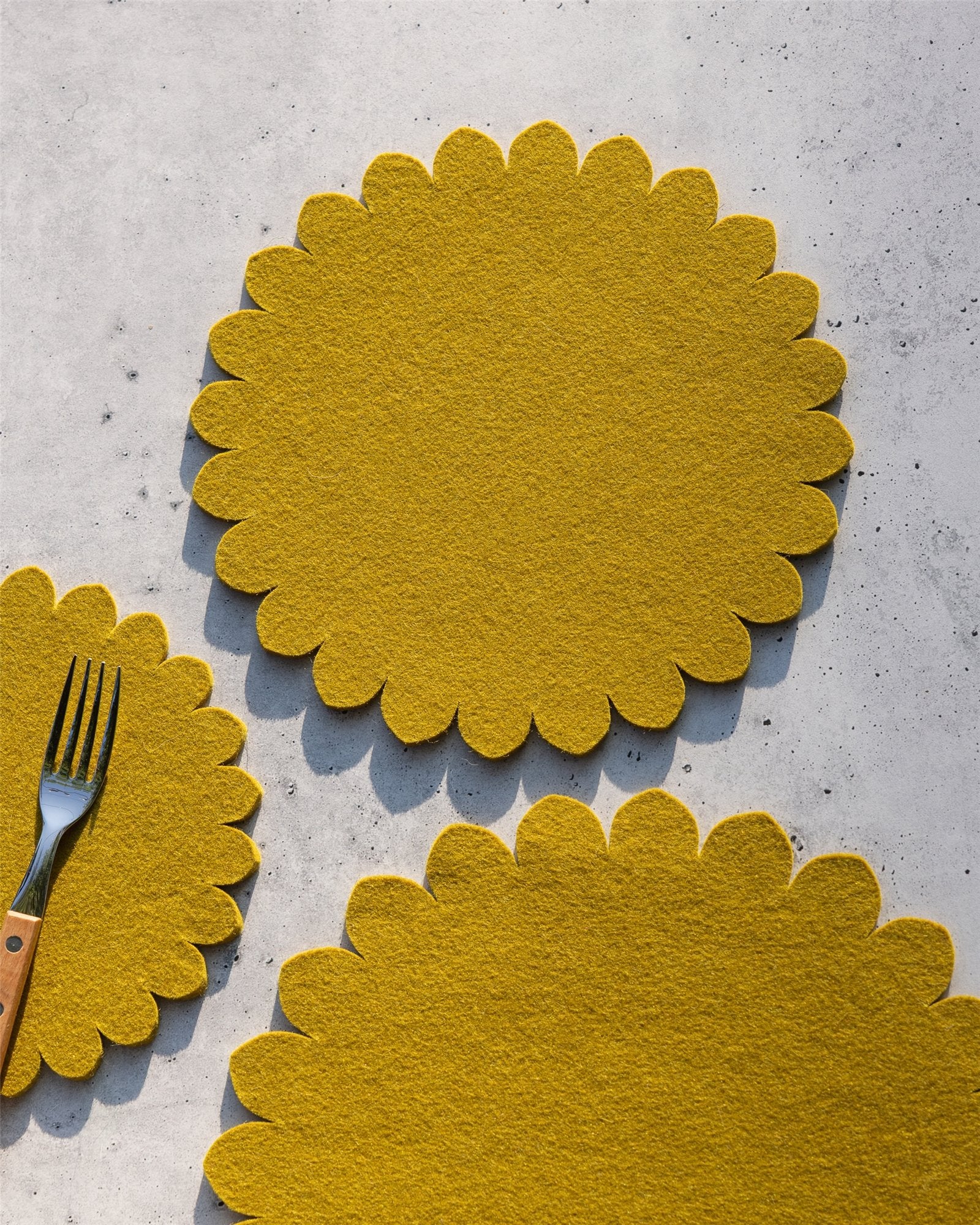 Three yellow, scalloped-edge graf lantz Merino wool trivets are arranged on a light speckled surface. A metal fork with a wooden handle rests on the smallest trivet in the bottom left corner.