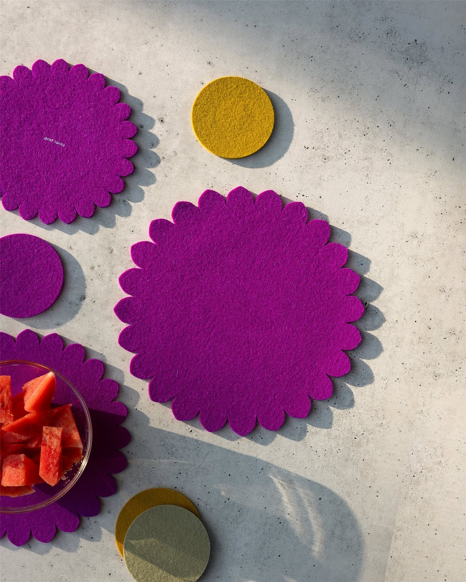 Graf lantz round scalloped magenta felt placemats and yellow coasters, inspired by a Japanese floral crest, are arranged on a light textured surface with a glass bowl of watermelon pieces in the lower left corner.