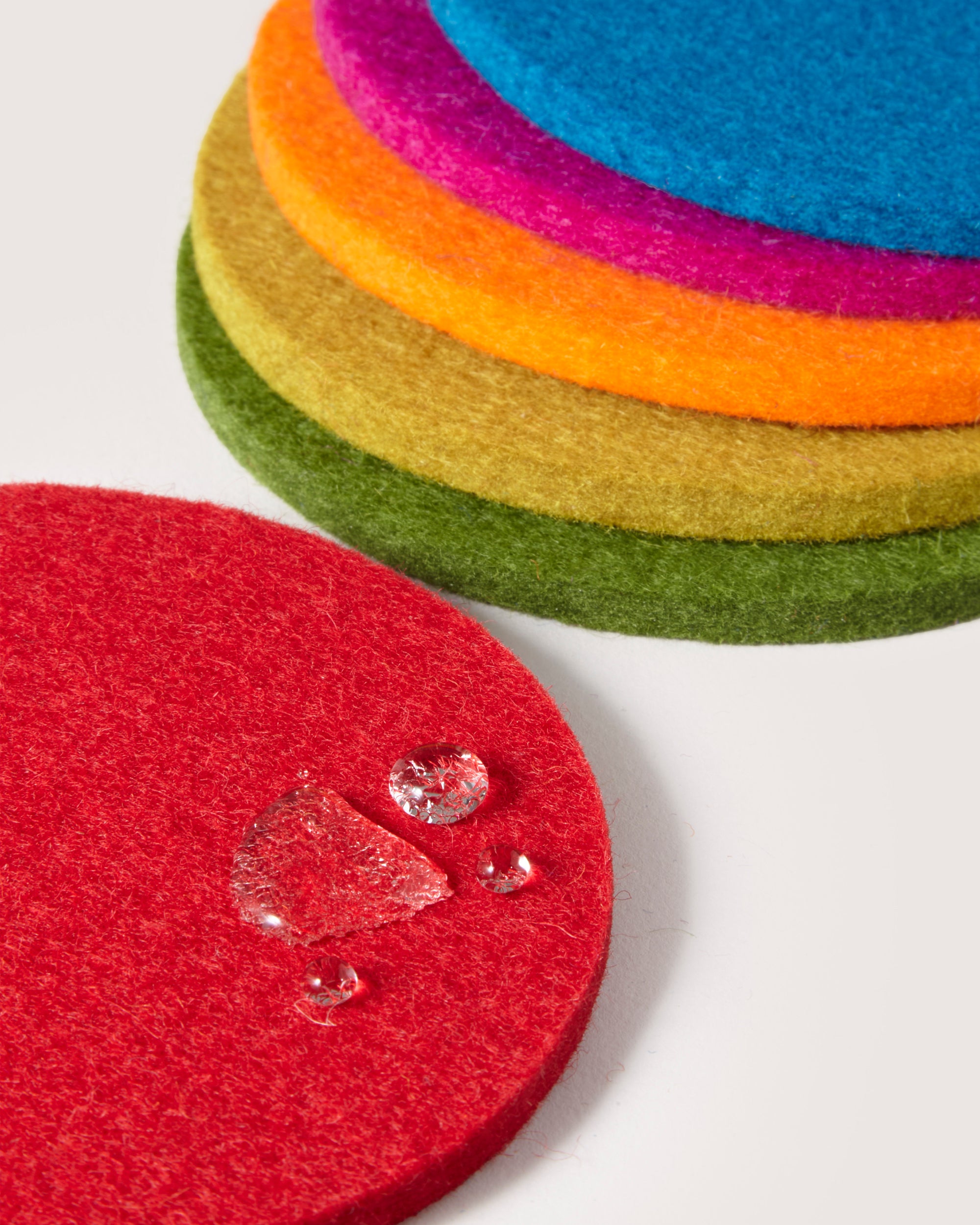 A stack of vibrant felt coasters by graf lantz, made from German Merino wool in red, orange, yellow, pink, and green. The foreground features a red coaster adorned with water droplets, highlighting its texture and absorbency.