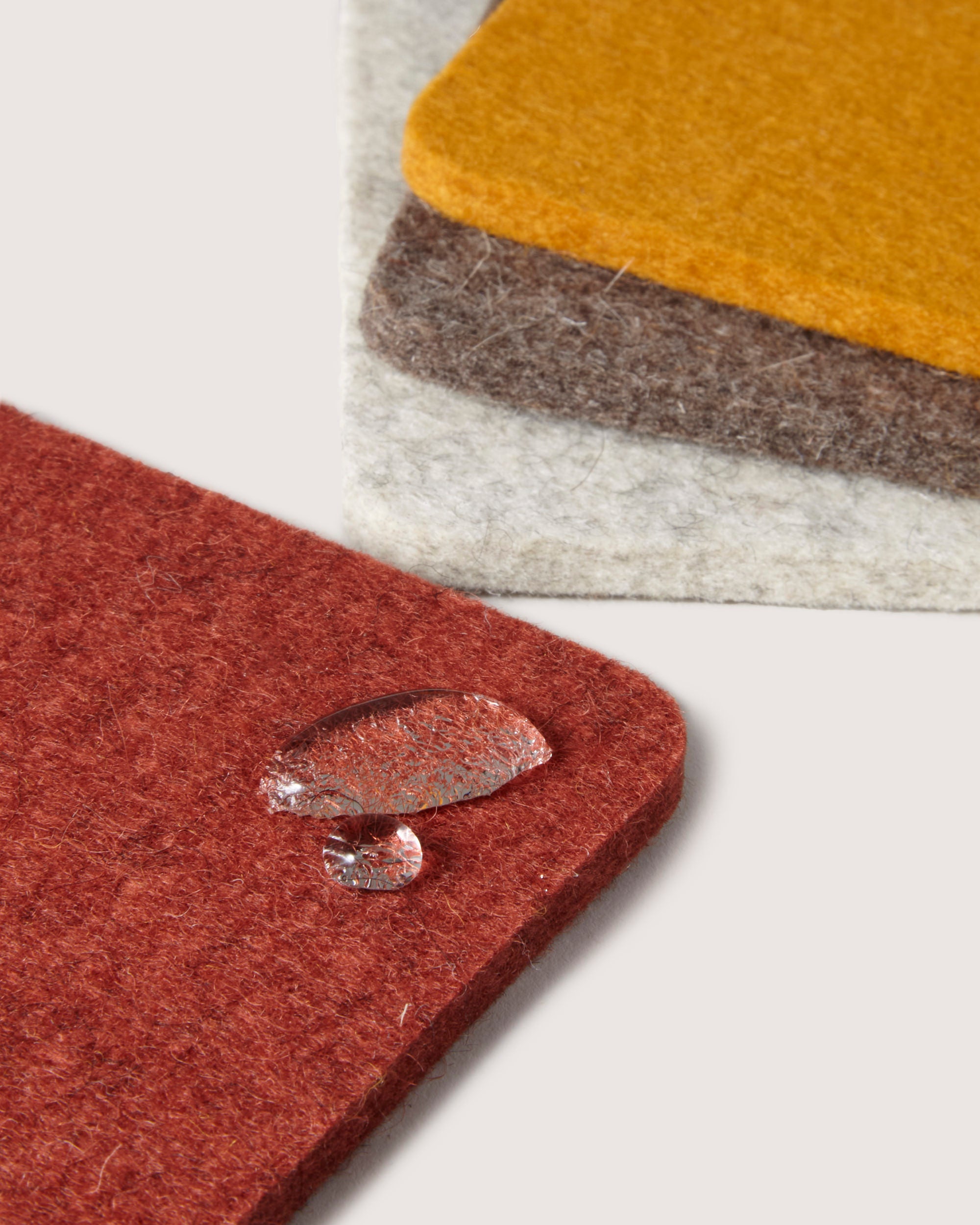 A fan-like stack of four graf lantz coasters made from German Merino wool in red, yellow, brown, and gray highlights their premium quality. A water droplet sits on the red coaster, emphasizing its water resistance.