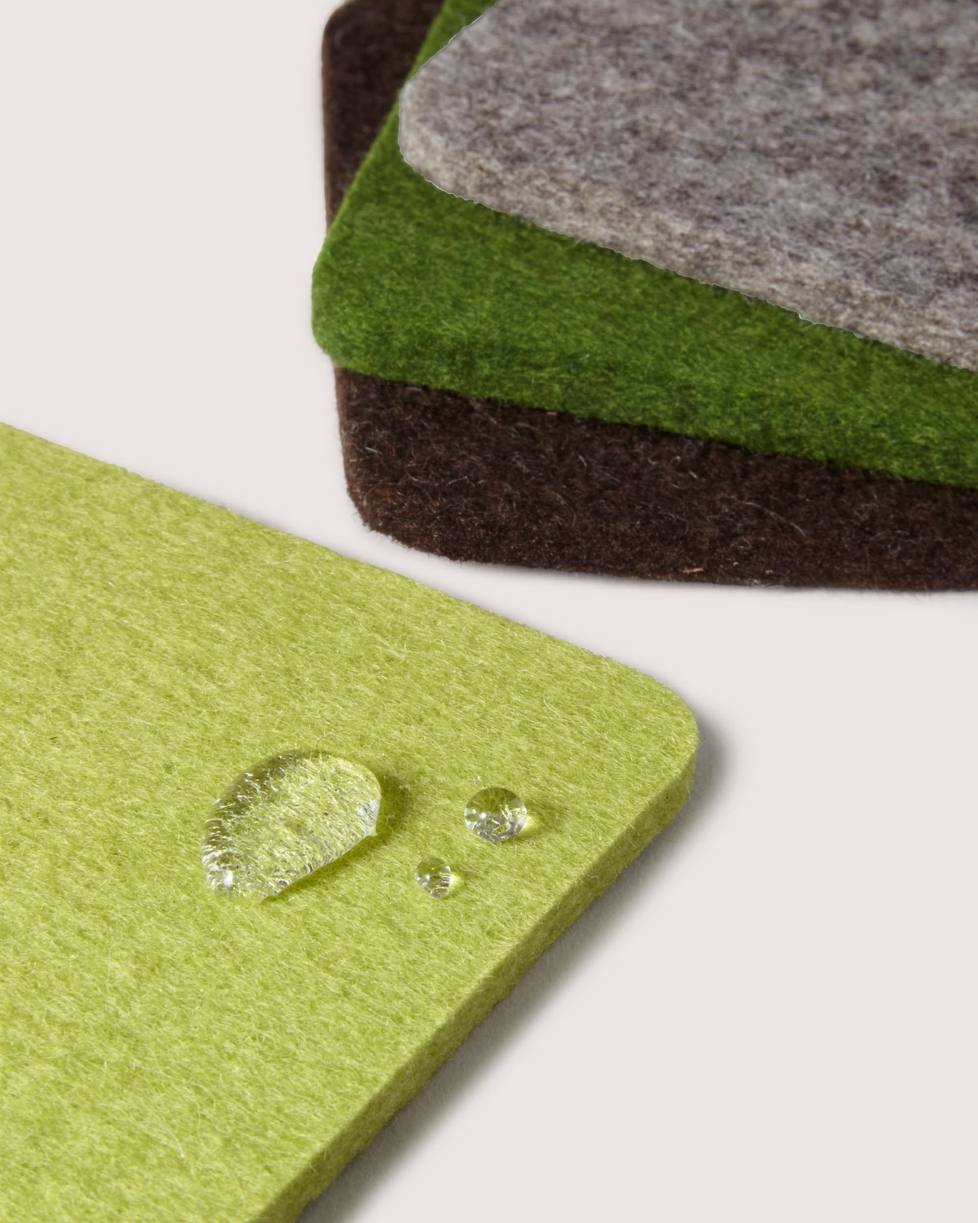 Close-up of four square coasters in lime green, green, brown, and gray crafted from Merino wool by graf lantz on a white surface. The lime green coaster features water droplets, highlighting its water-resistant design inspired by German heritage.