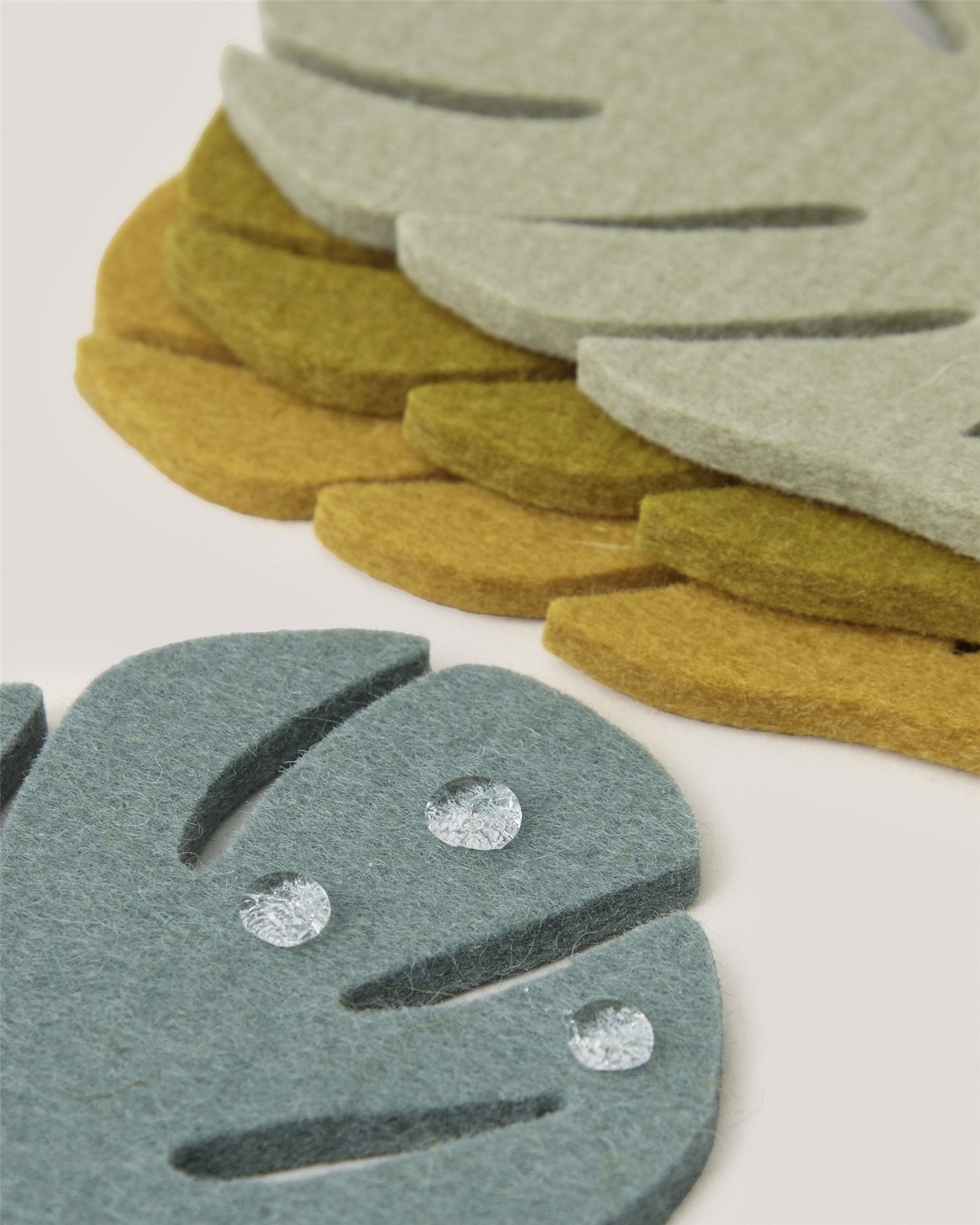Close-up of layered Merino wool Graf Lantz Monstera coasters in green, mustard yellow, and gray-blue—a stylish, sustainable home accent. The front coaster features three visible water droplets on its surface.
