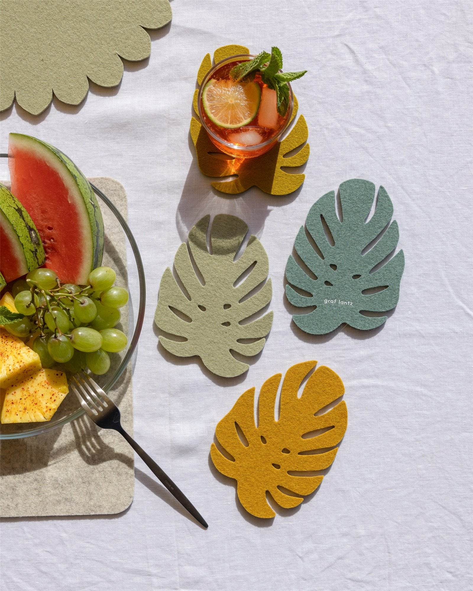 A white tablecloth holds a plate of watermelon, pineapple, and grapes beside a fork. Three Graf Lantz Monstera leaf coasters made of Merino wool and a drink with orange and mint complete the setting.
