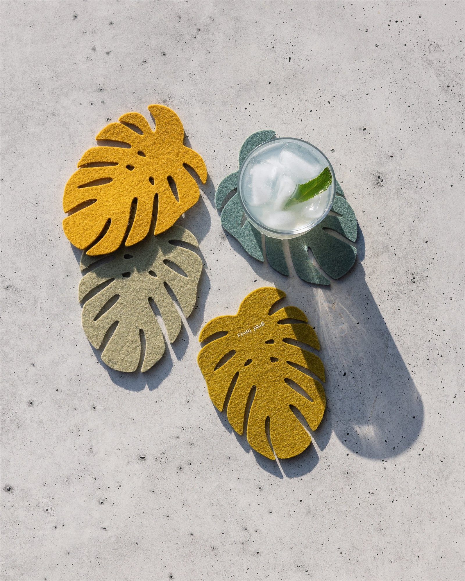 Three Monstera coasters by Graf Lantz, made from premium Merino wool in yellow and green, are displayed on a light gray speckled surface. One holds a glass of iced water with a leaf garnish—ideal for sustainable home decor.