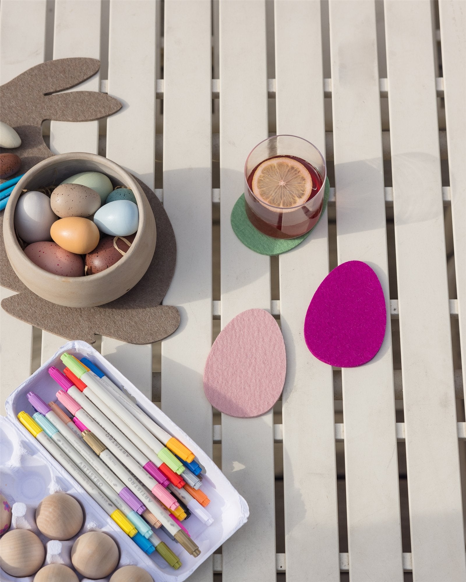 A Graf Lantz table setup features a bowl of colorful eggs, colored pens, and pastel felt egg shapes. A lemon glass rests on an egg coaster with Merino wool accents. Perfectly organized for crafting or decoration.