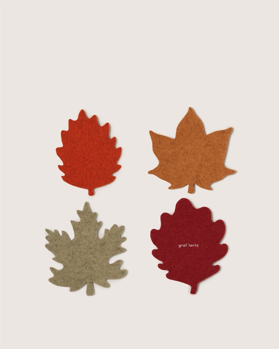 Four felt coasters by Graf Lantz, each shaped like a different autumn leaf, arranged in a grid. The coasters come in red, orange, tan, and light brown; the red coaster features the Graf Lantz logo.