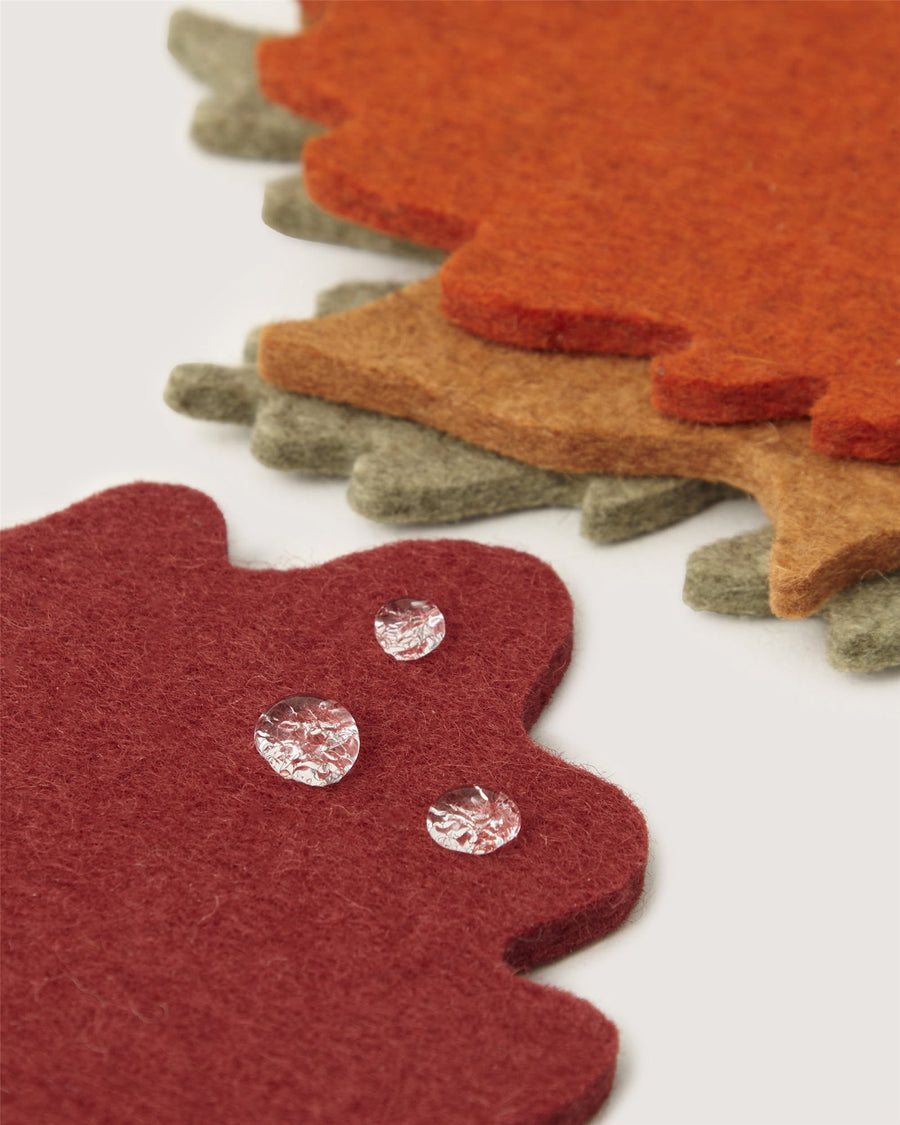 Three textured felt coasters by Graf Lantz in red, orange, and brown are layered with jagged edges. Three water droplets rest on the red coaster in the foreground.
