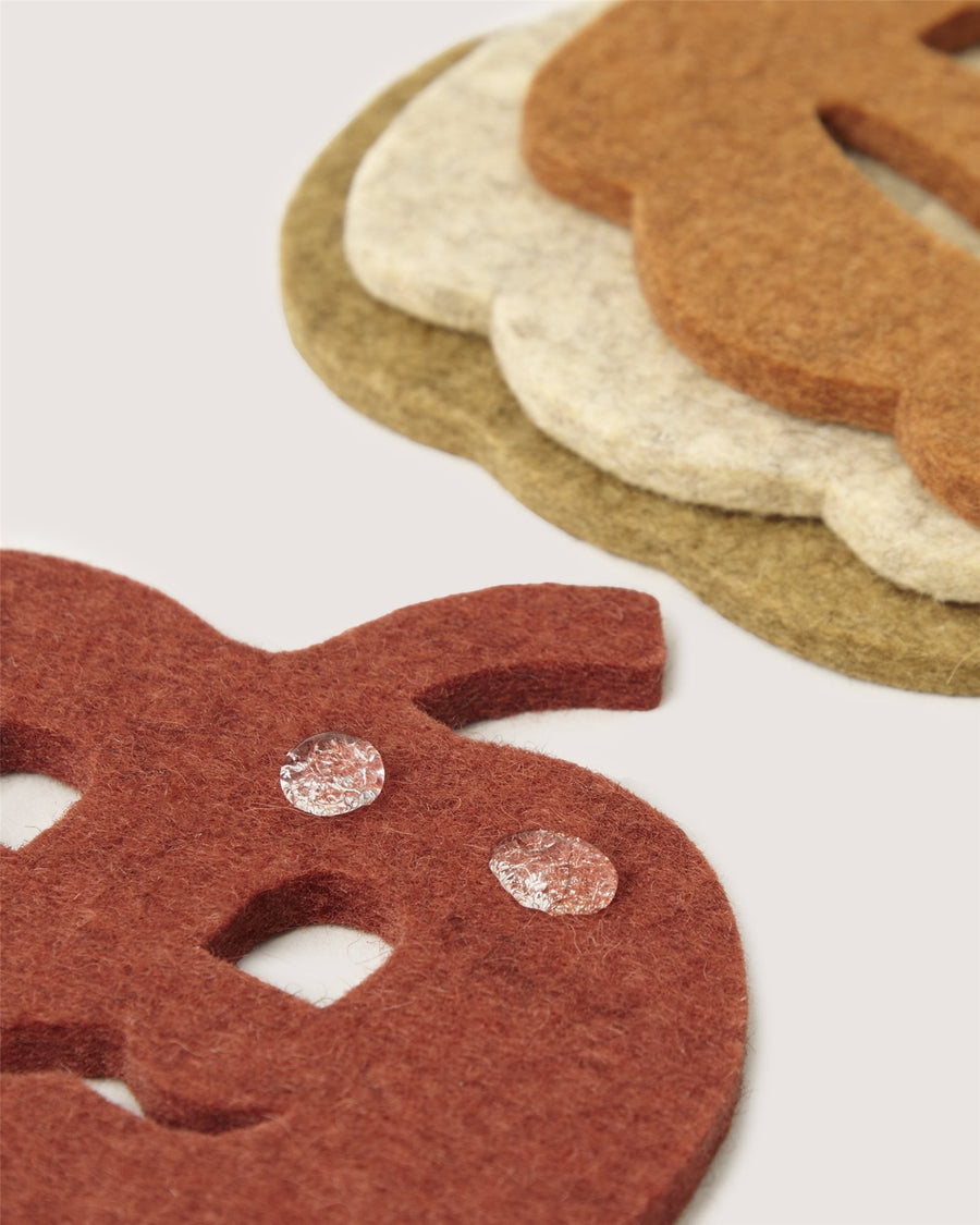 Three layered Graf Lantz felt cutouts in pumpkin and leaf shapes are arranged on a light surface. Two small, round water droplets rest on the orange pumpkin-shaped felt in the foreground.