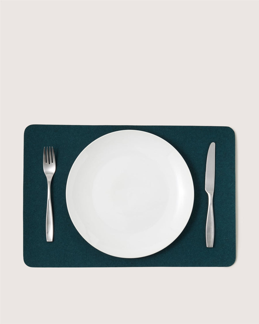 A white plate is centered on a Graf Lantz dark green rectangular placemat, with a silver fork on the left and a silver knife on the right, all placed on a light-colored surface.