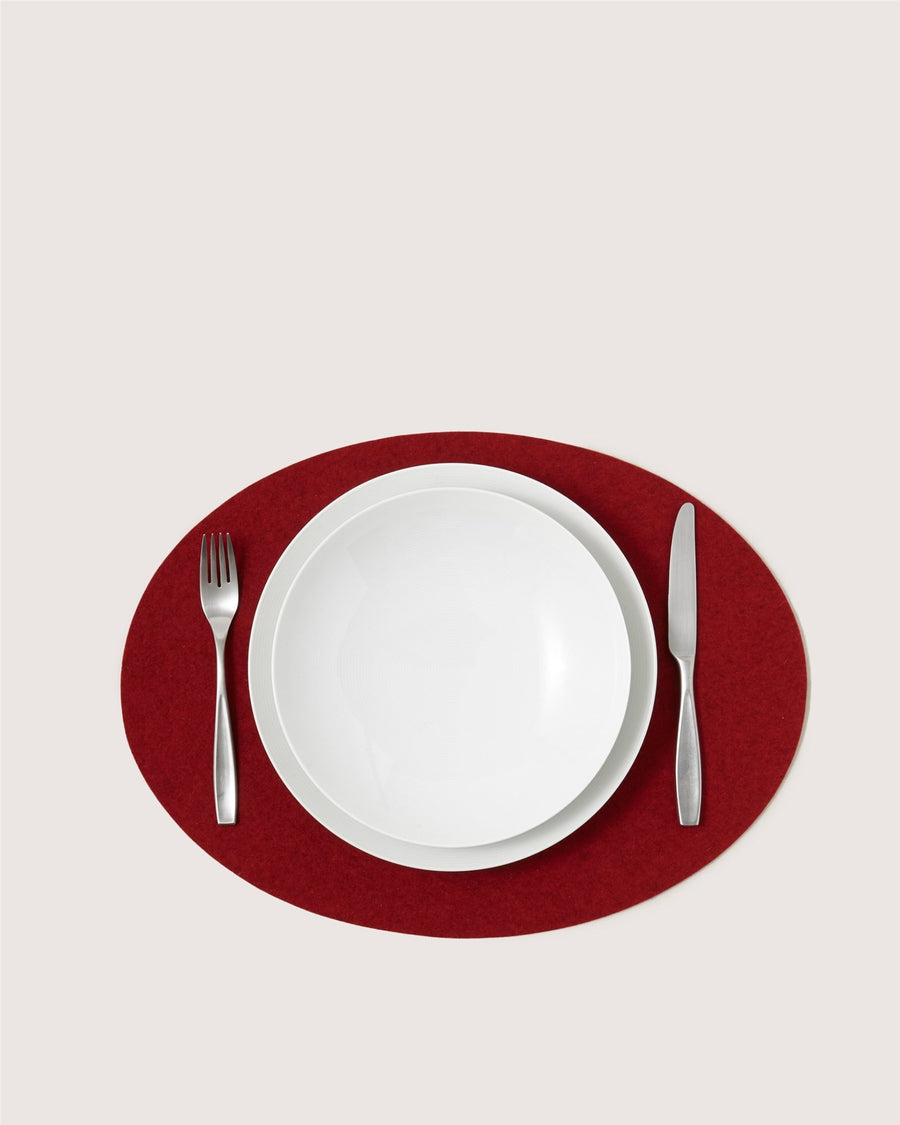 A white plate and bowl from Graf Lantz rest on a red oval placemat, fork to the left and knife to the right, all set against a light background.