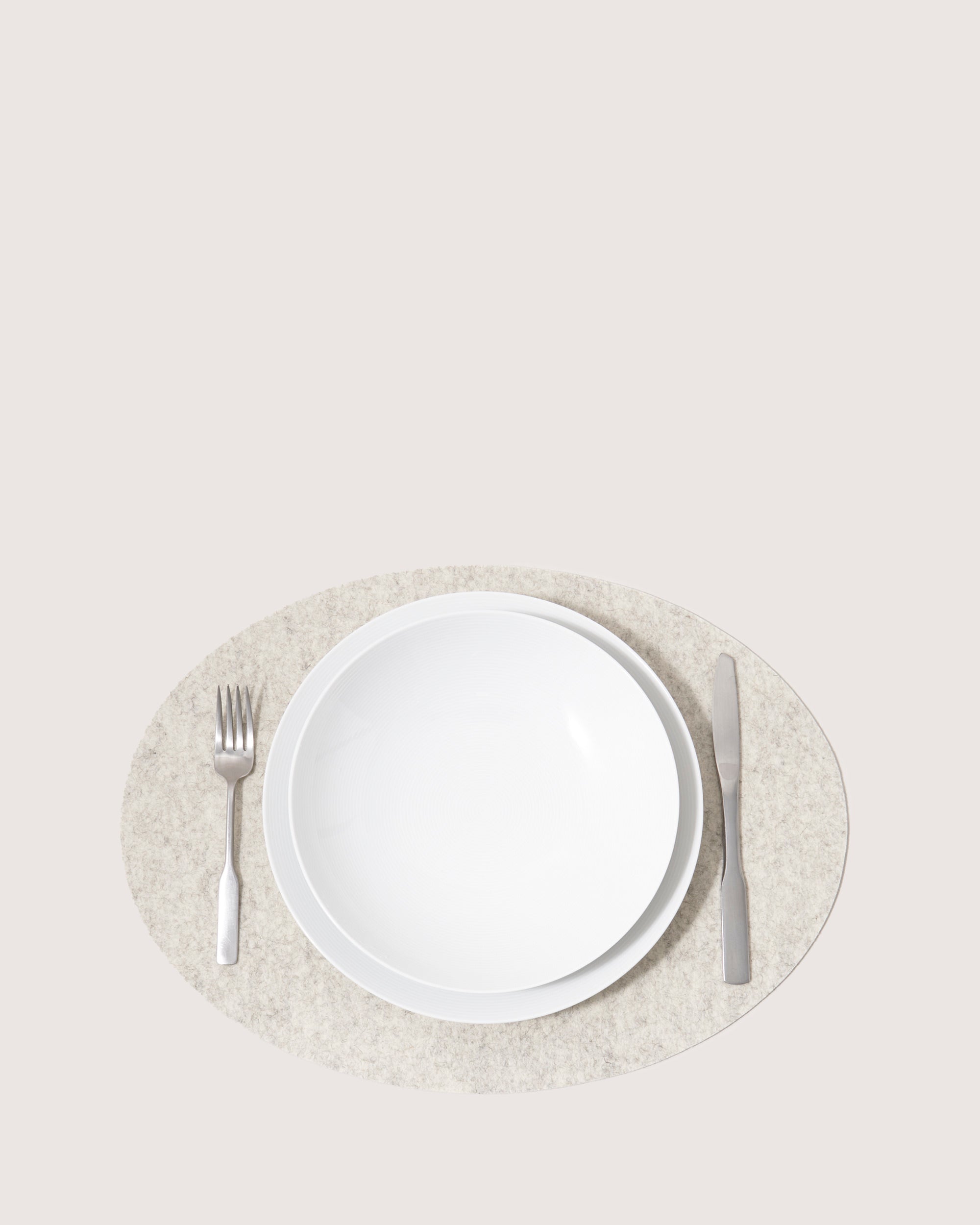 A white plate sits on a sophisticated graf lantz Merino Wool Placemat in light gray. To the left lies a stainless steel fork, and to the right, a knife. The light beige background accentuates this moisture-wicking and heat-resistant setups elegant allure.