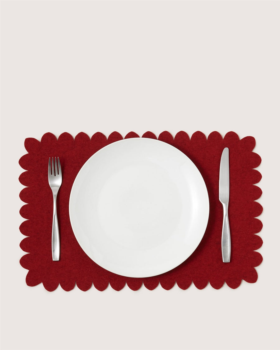 A Graf Lantz white plate rests on a red scalloped placemat, with a fork to the left and a knife to the right, set against a light background.