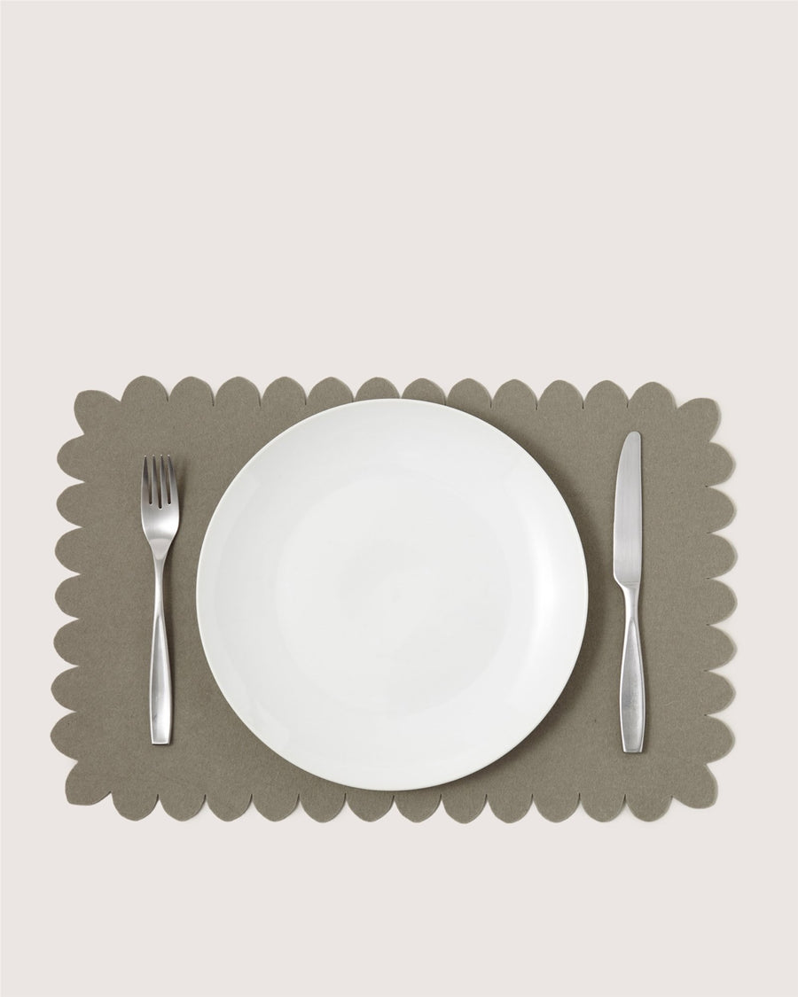 A white plate sits on a Graf Lantz gray scalloped placemat, flanked by a stainless steel fork on the left and knife on the right, all placed against a light background.