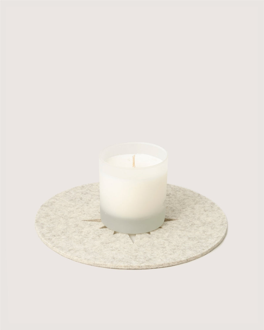 A white candle in a frosted glass holder by Graf Lantz rests on a round, textured beige coaster against a plain light background.