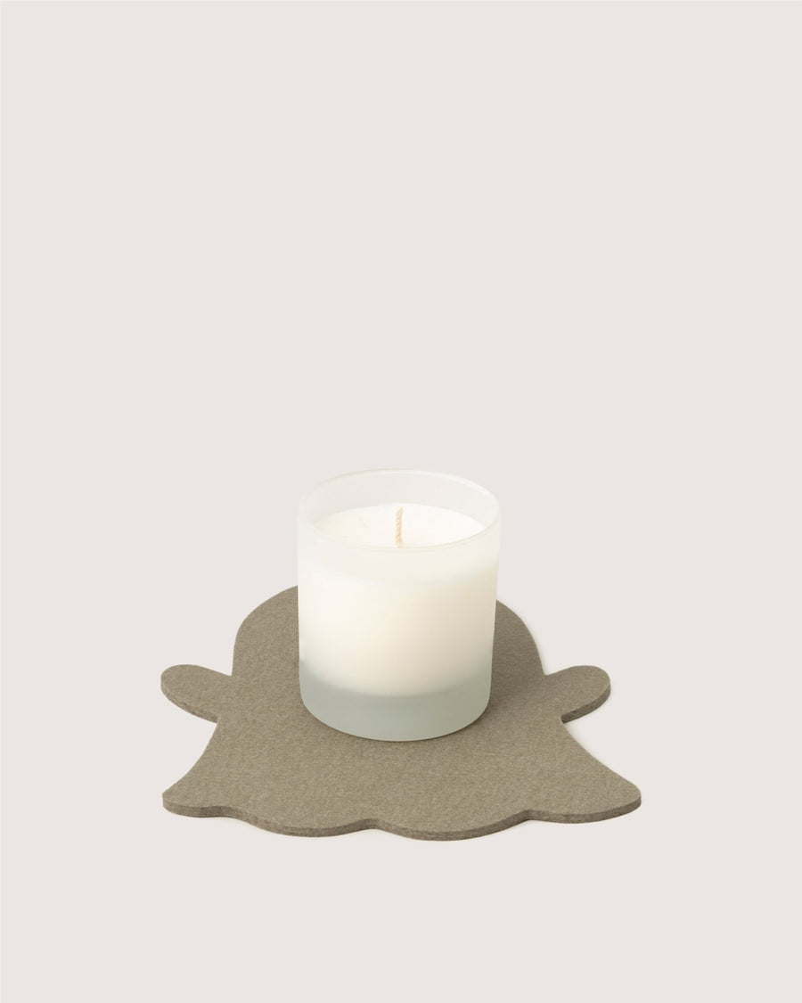 A white candle in a frosted glass holder by Graf Lantz rests on a gray, ghost-shaped coaster against a plain, light background.