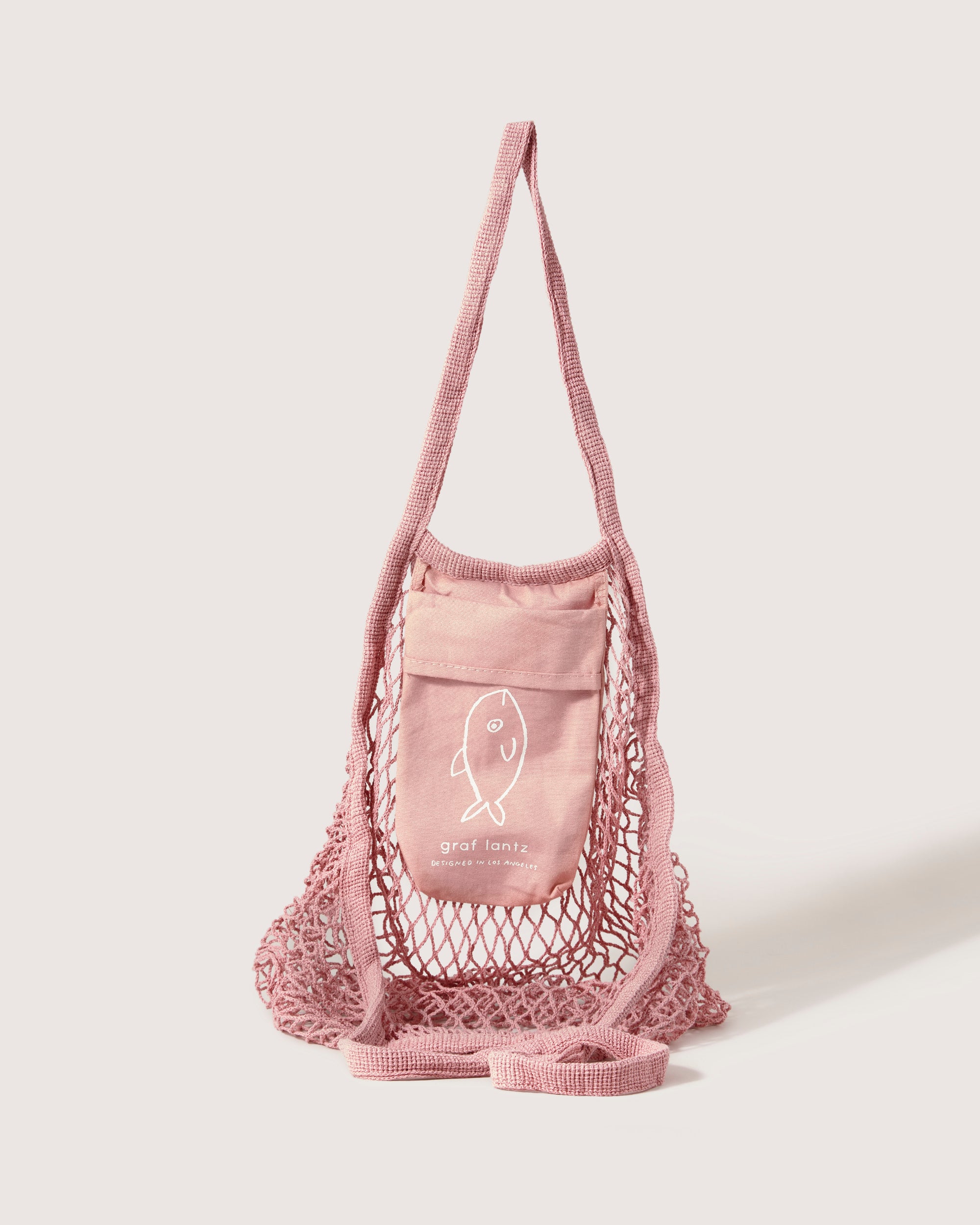 Graf Lantz presents a pink mesh bag featuring an expandable design with a long strap. It has a pocket adorned with a simple fish graphic and text, crafted in a net market tote style and elegantly showcased against a plain background.