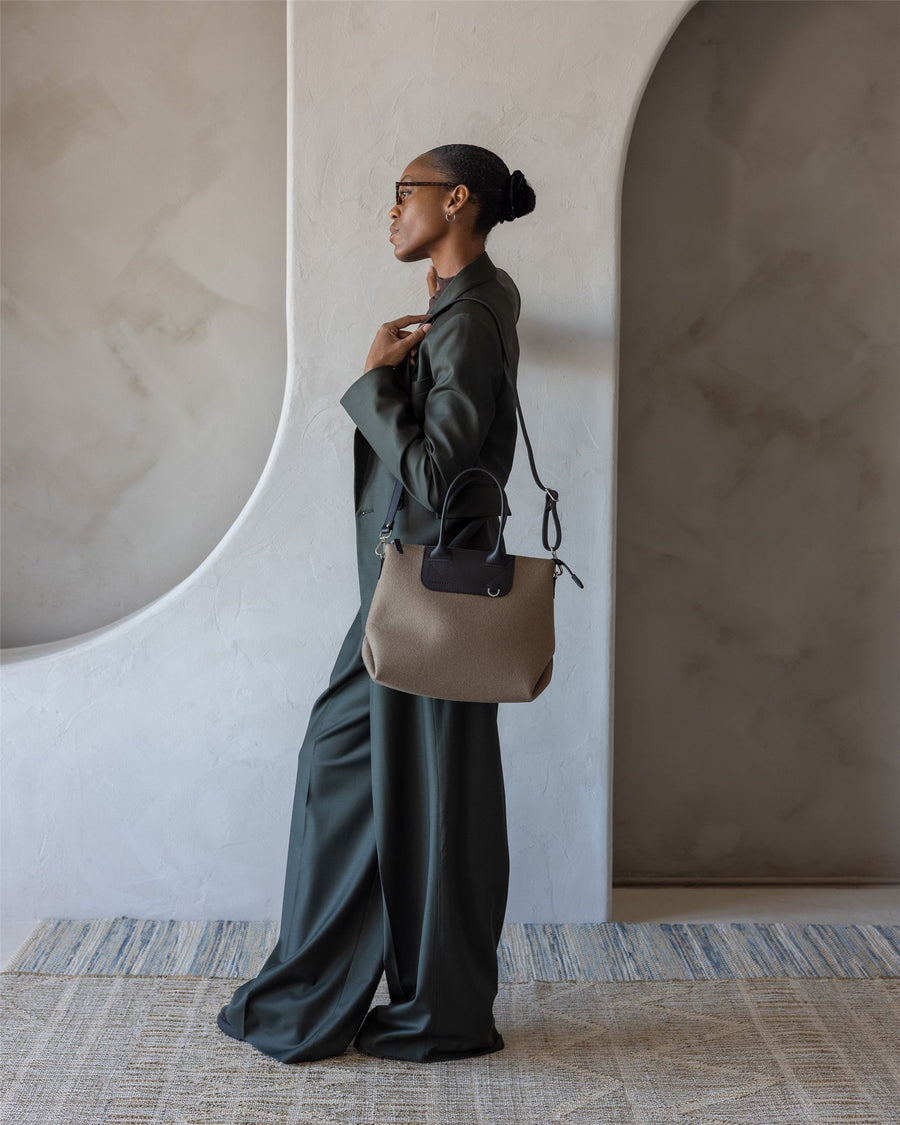 A woman in a dark green pantsuit stands sideways against a light-colored textured wall, holding a taupe Graf Lantz handbag over her shoulder. She wears glasses and has her hair neatly pulled back in a bun.