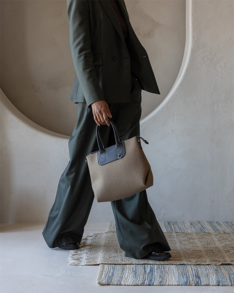 A person in a dark green suit walks indoors on a rug, holding a brown and black Graf Lantz handbag. The curved, textured wall niche is in the background. The person’s face is not visible.