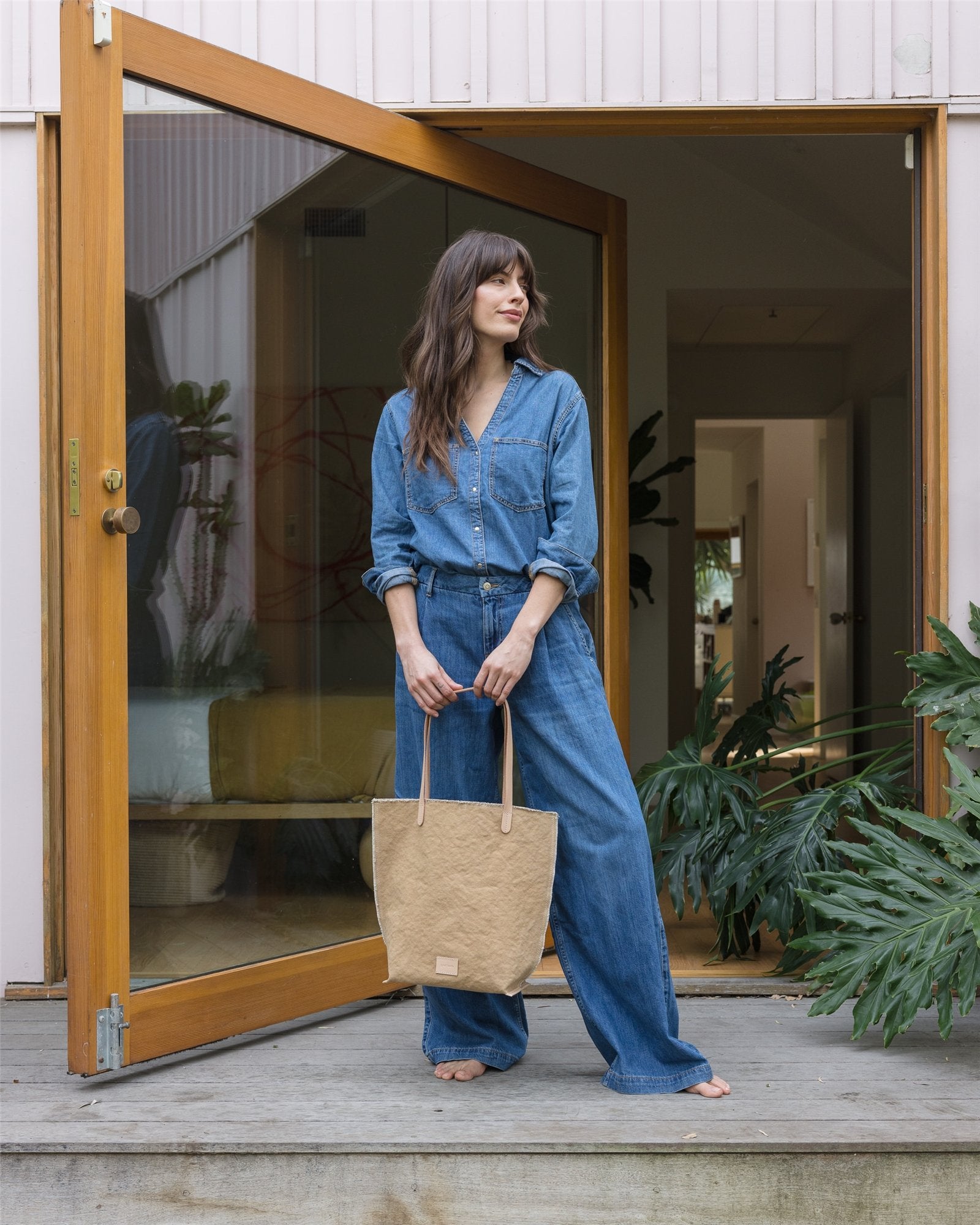 A person in a denim jacket and jeans stands barefoot at an open glass door, holding a Graf Lantz beige canvas tote with leather handles. They glance to the side, with plants visible in the foreground and a cozy interior featuring a yellow sofa in the background.