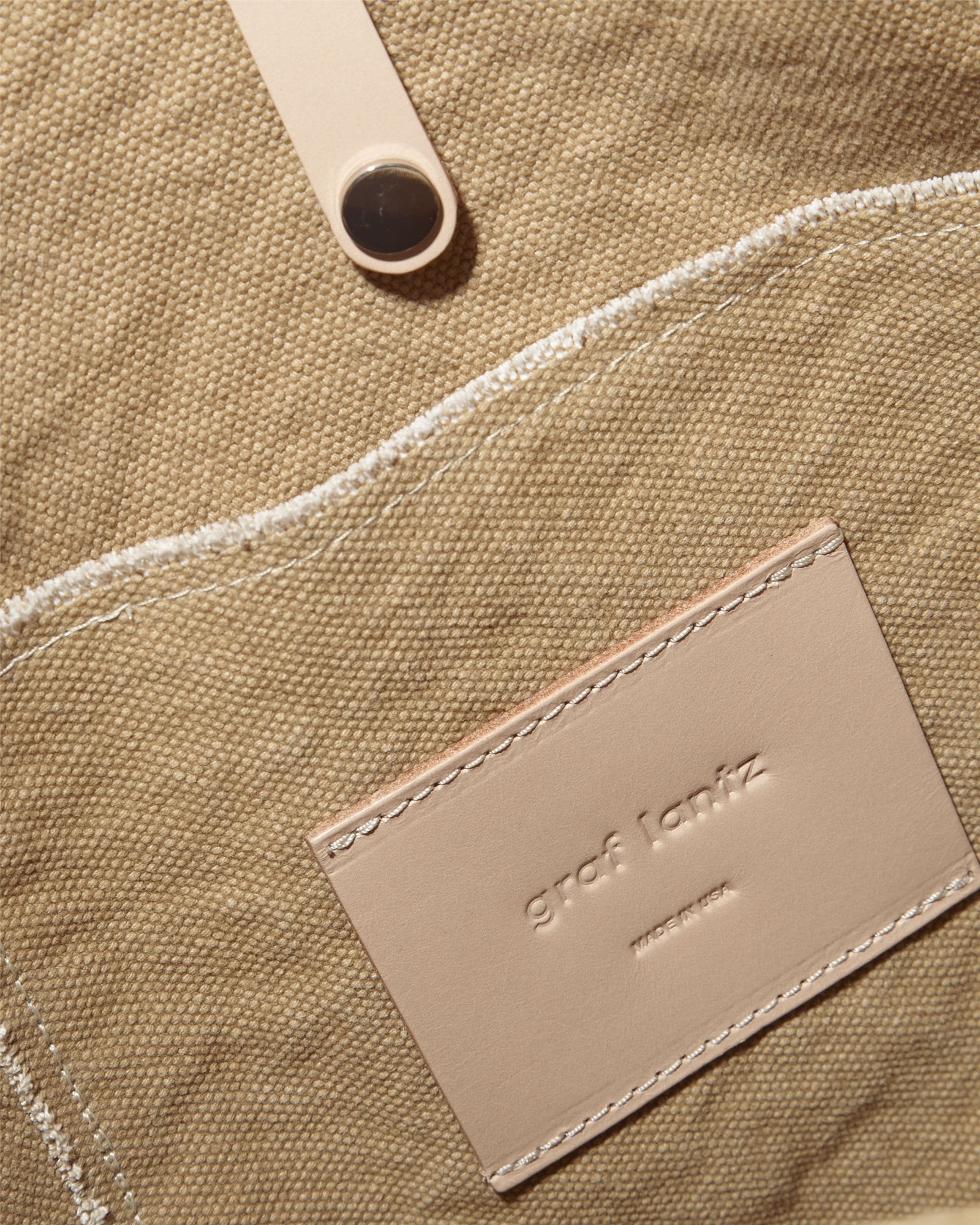 A close-up of the Hana Canvas Boat Bag shows a textured beige fabric surface with a rectangular label that has Graf Lantz embossed in small letters. The label is sewn on with white stitching, features a button detail above it, and has genuine leather handles enhancing its design.