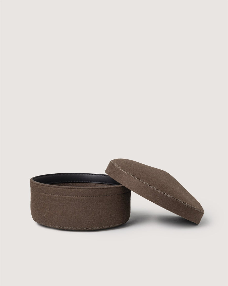 A round, brown fabric container by Graf Lantz sits empty on a light background, its matching lid placed slightly tilted beside it.