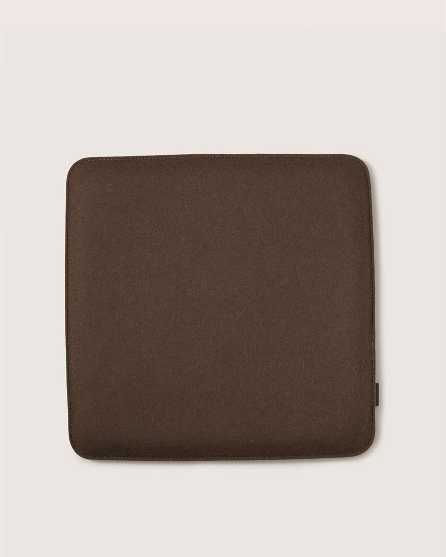 A square, dark brown Graf Lantz cushion with rounded corners sits on a light beige background. Its surface looks soft and smooth.