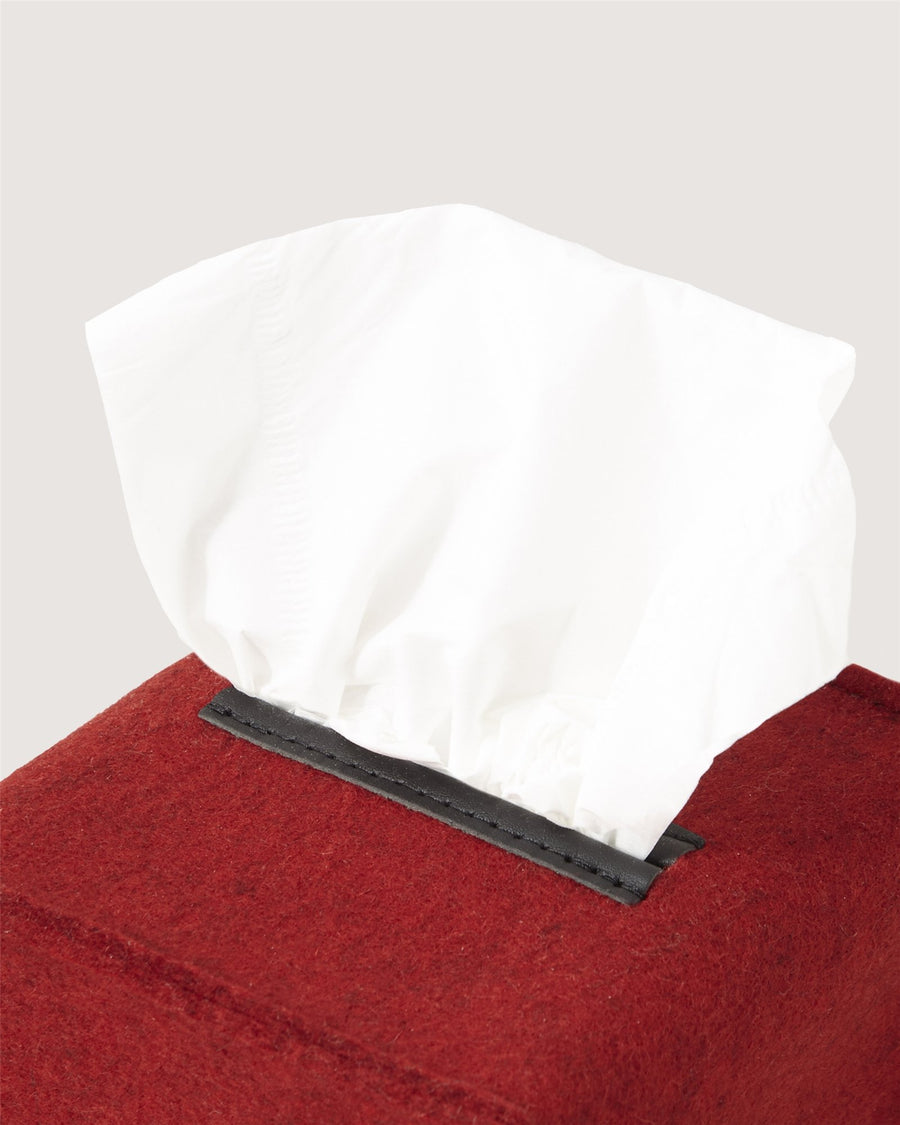 A close-up of a white tissue protruding from a rectangular, red fabric Graf Lantz tissue box with a black plastic opening.