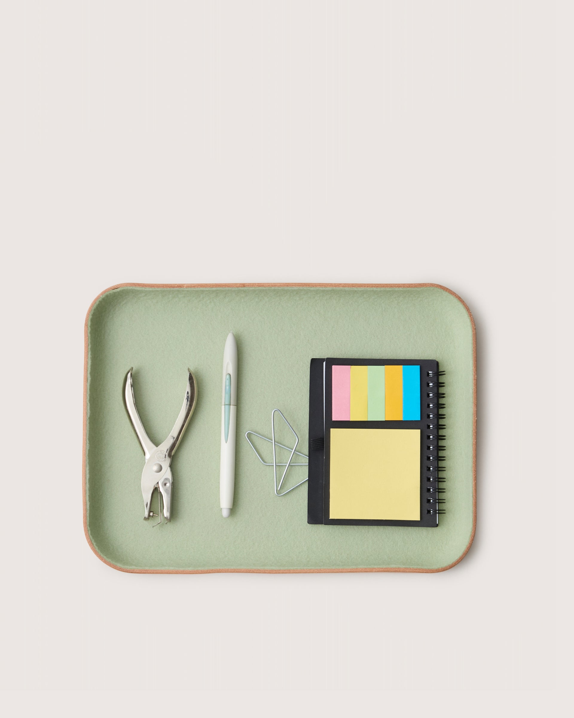 Green tray with office supplies including a hole punch, pen, paper clips, and a small notebook on a light gray background.