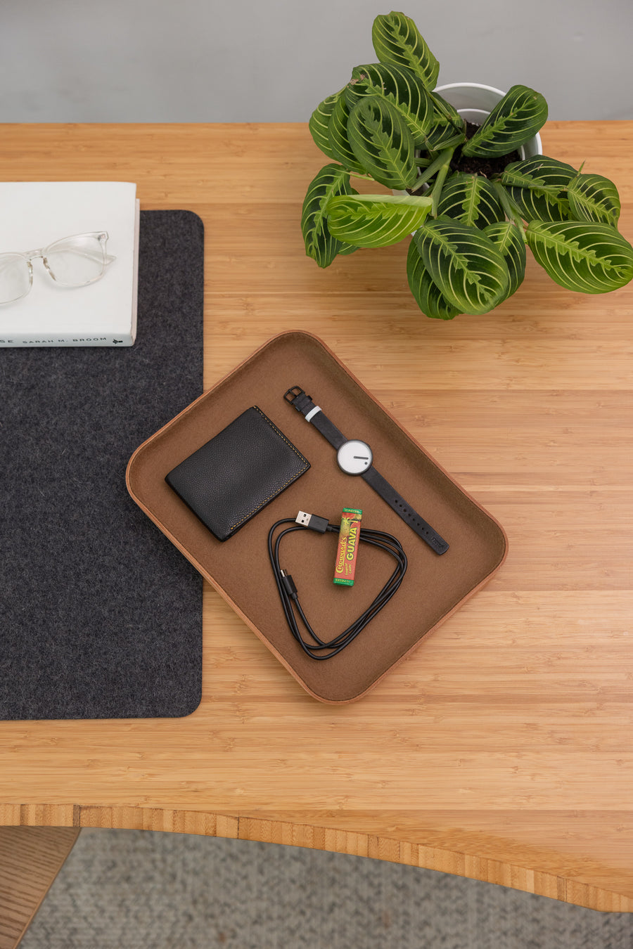 A wooden desk with a plant, a book topped with eyeglasses, and a graf lantz Kawabon Tray holding a black wallet, smartwatch, USB cable, and chewing gum.
