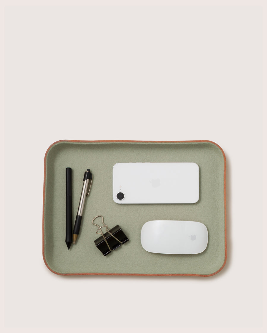 A green graf lantz Kawabon Tray holds a black pen, a black and silver pen, two black binder clips, a white smartphone, and a white wireless mouse, all arranged neatly on a light background.