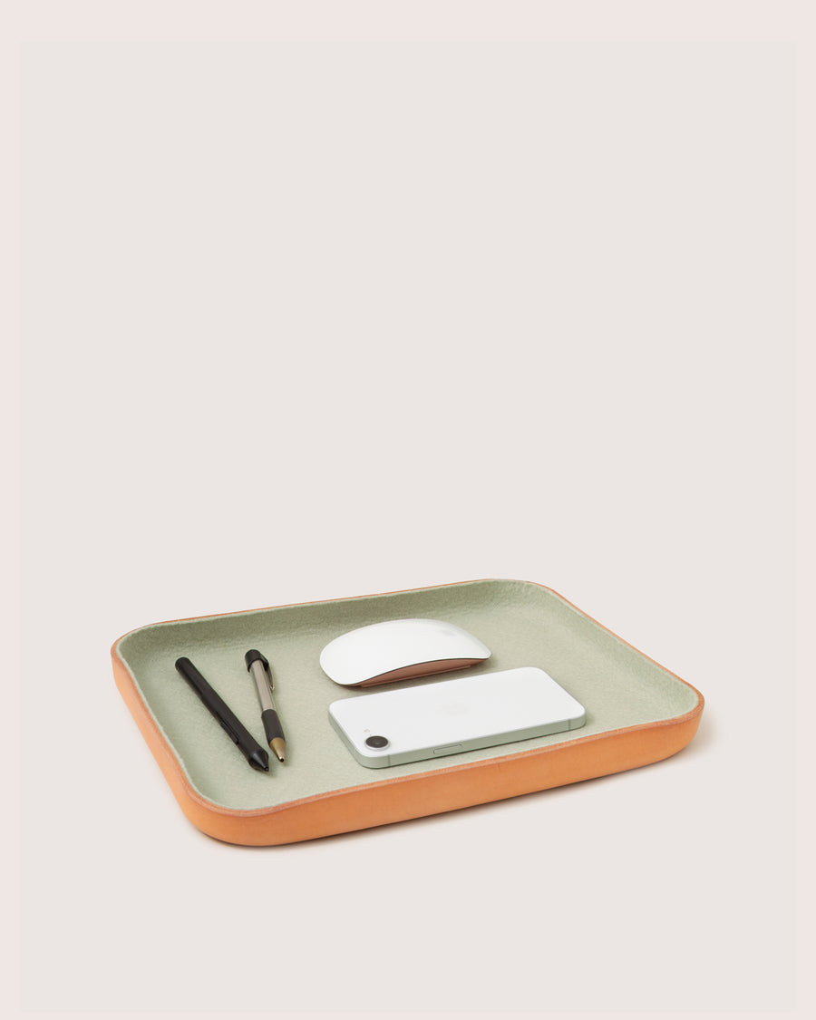 A rectangular graf lantz Kawabon Tray with a green Merino wool interior and tan edges holds two pens, a white wireless mouse, and a white smartphone on a light surface against a plain background.