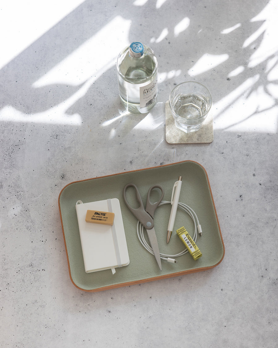 A graf lantz catchall tray holding a notebook, eraser, scissors, pen, charging cable, and candy sits on a gray surface. Nearby are a glass of water on a coaster and sparkling water with leafy shadows across the table.