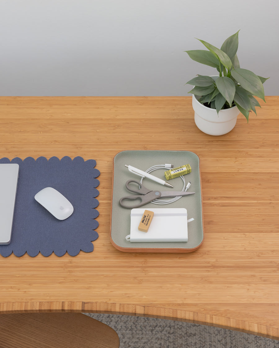 A tidy wooden desk with a mouse, blue scalloped graf lantz Merino wool mat, gray graf lantz Kawabon Tray holding scissors, pens, cords, USB drive, eraser, and a potted plant in the corner against a light wall.